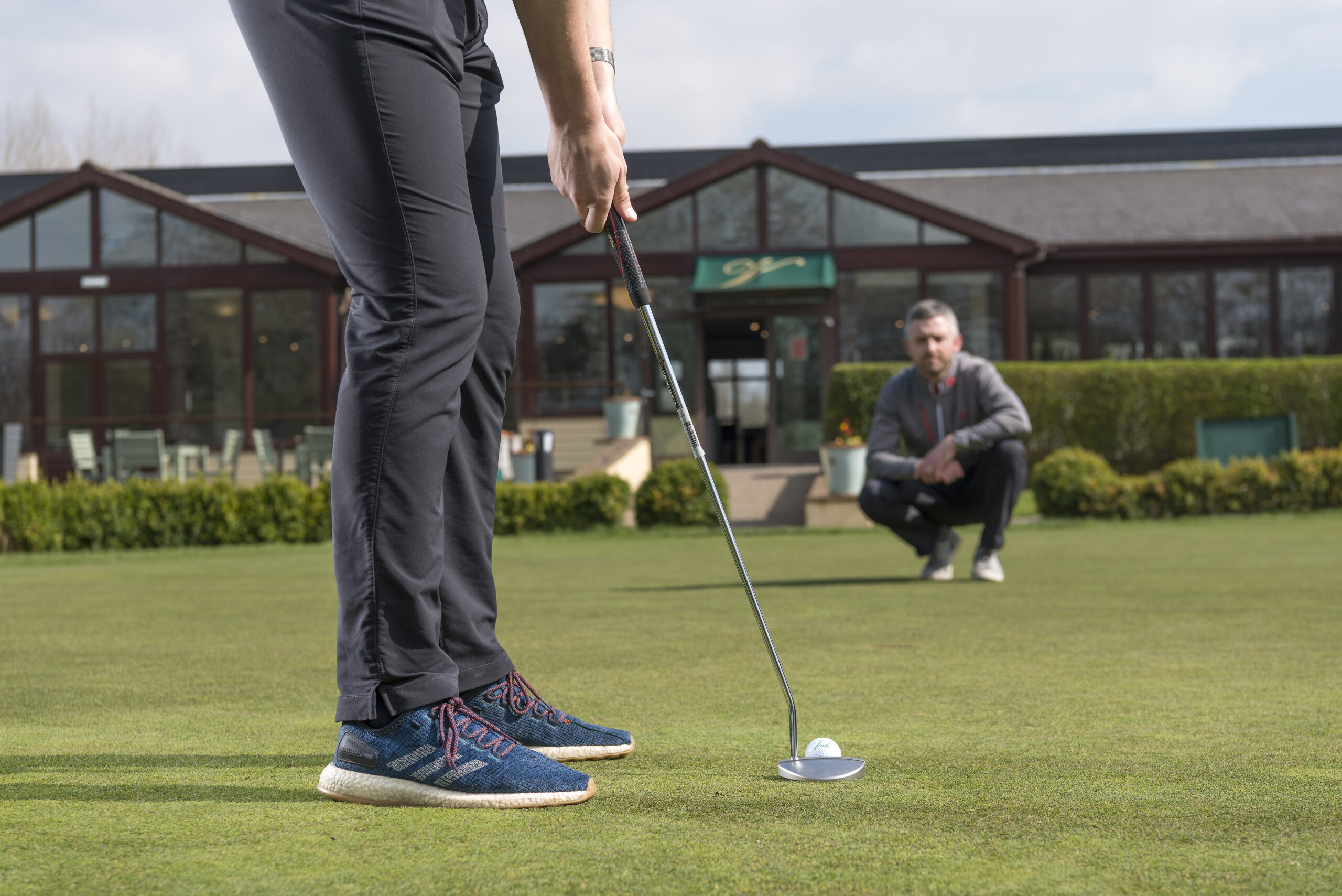 Person in sneakers prepares to putt a golf ball on a sunny day, while another person crouches in the background on the green.