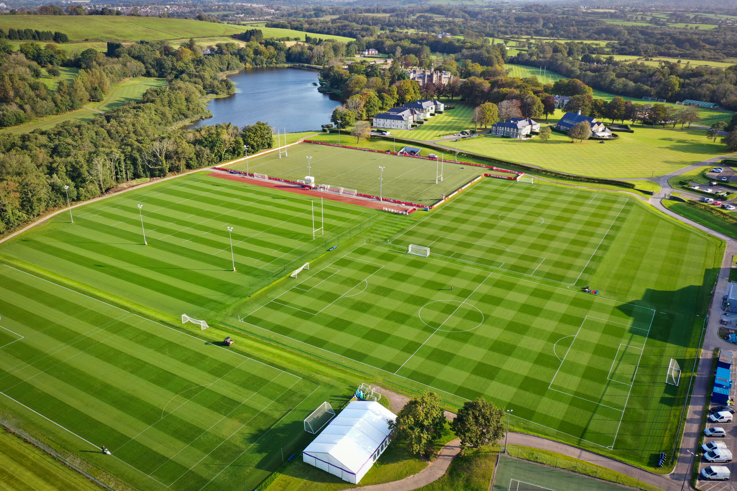 Aerial view of a large sports complex featuring multiple well-maintained soccer fields, surrounded by trees and a lake in the background.