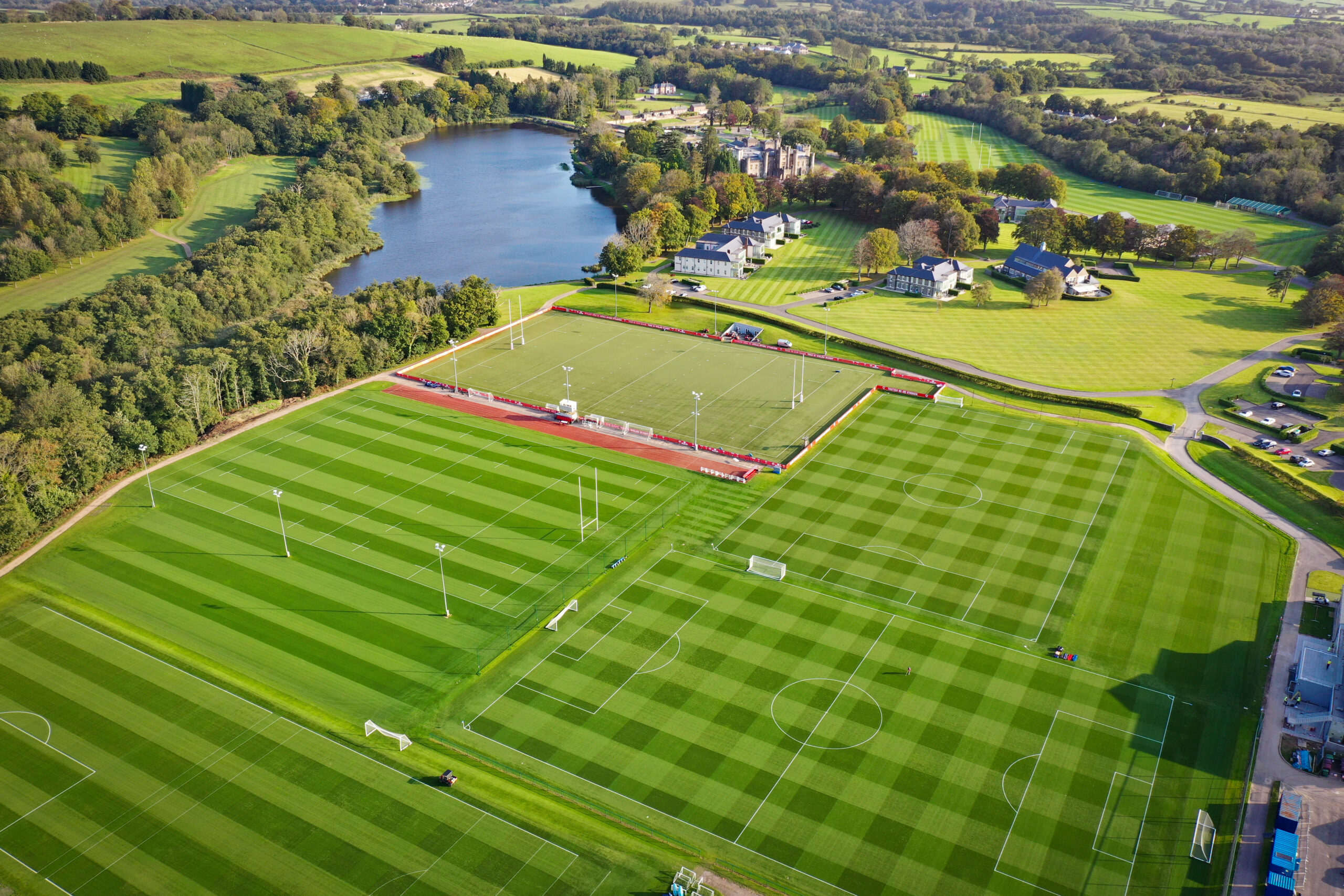 Aerial view of a large sports complex featuring multiple green soccer fields, bordered by trees and a lake, with buildings in the background.