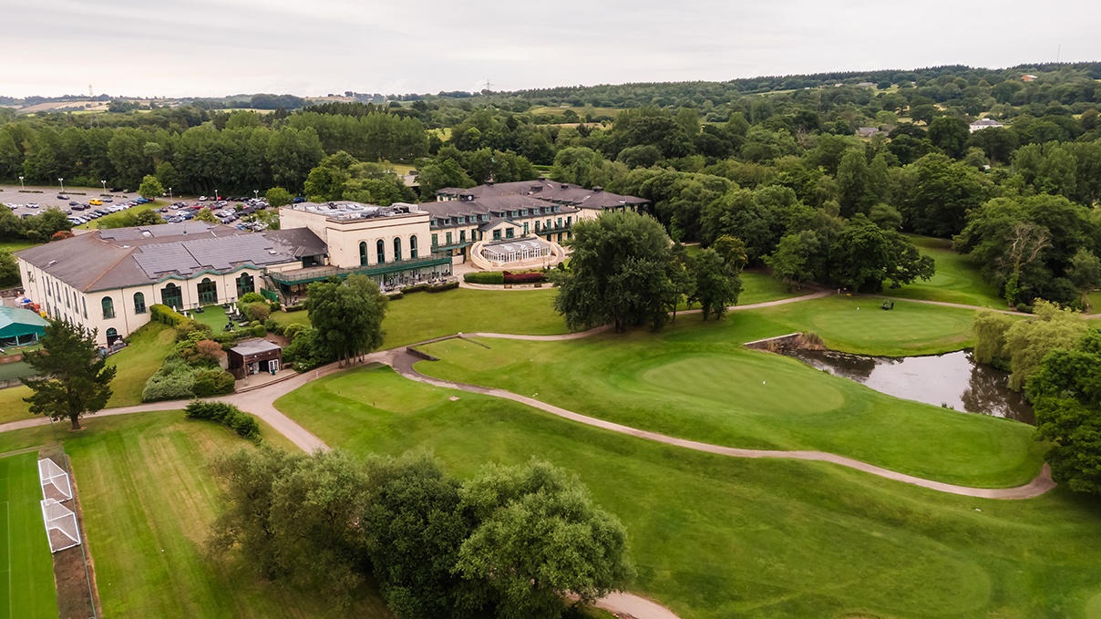 Aerial view of a golf course with a large clubhouse surrounded by trees and lush greenery. Paths and a small pond are visible on the grounds.