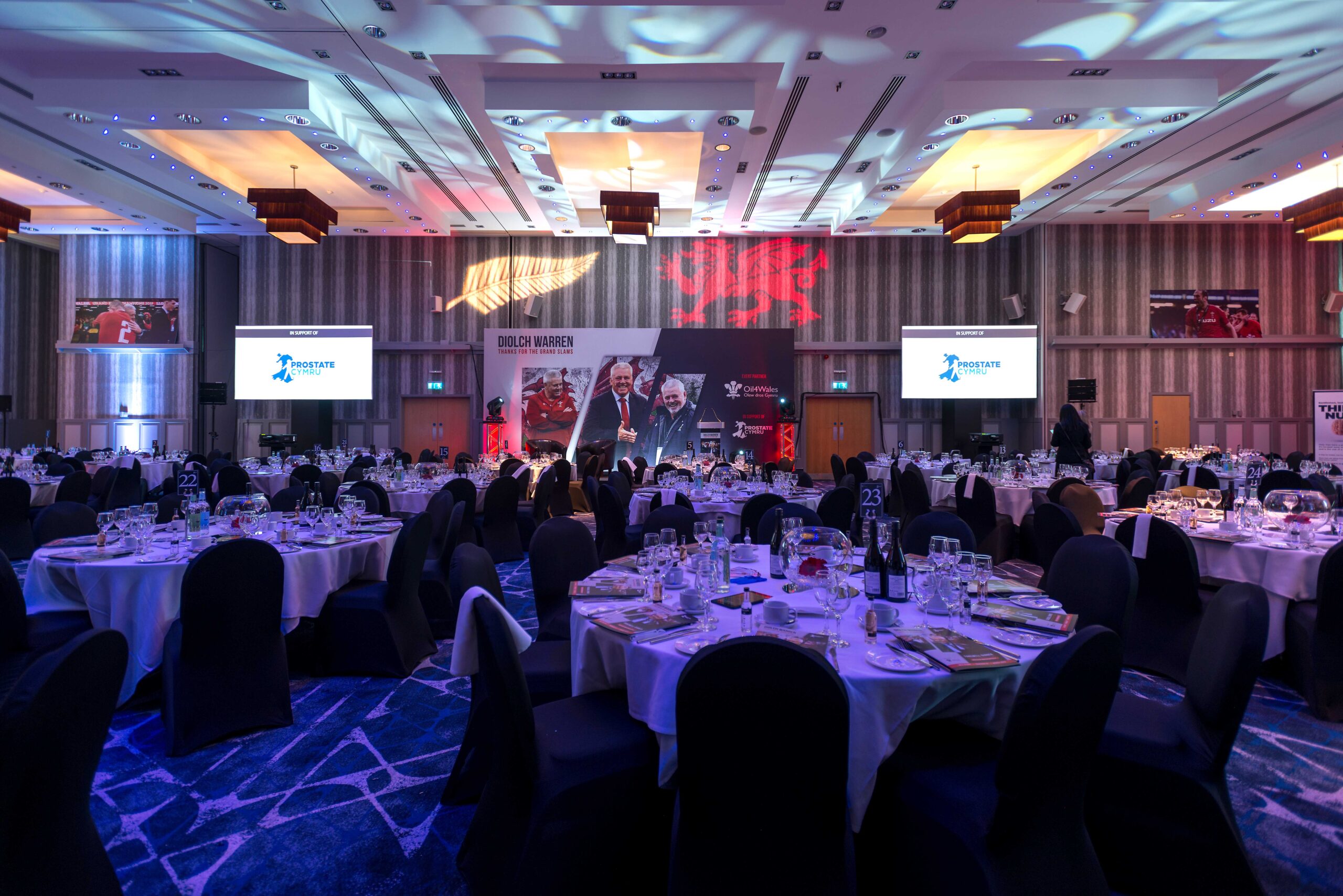 A conference room set up with round tables covered in white tablecloths, black chairs, and projection screens displaying a presentation logo.