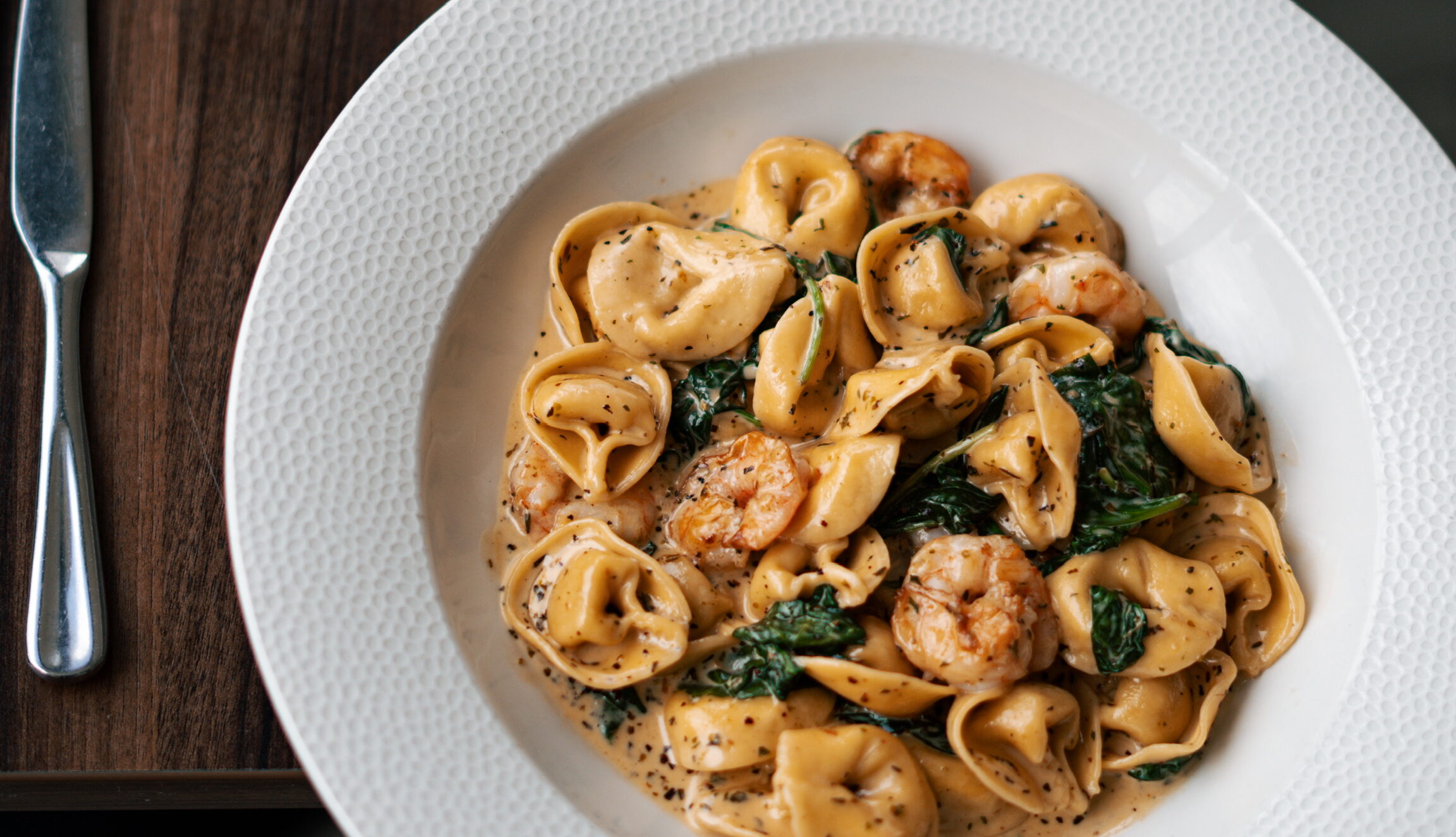 A white plate filled with creamy tortellini, shrimp, and wilted greens on a wooden table next to a knife.