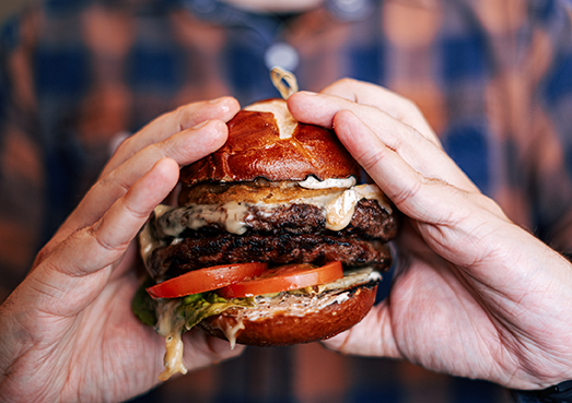 A person holds a large burger with lettuce, tomato, cheese, and a pretzel bun.