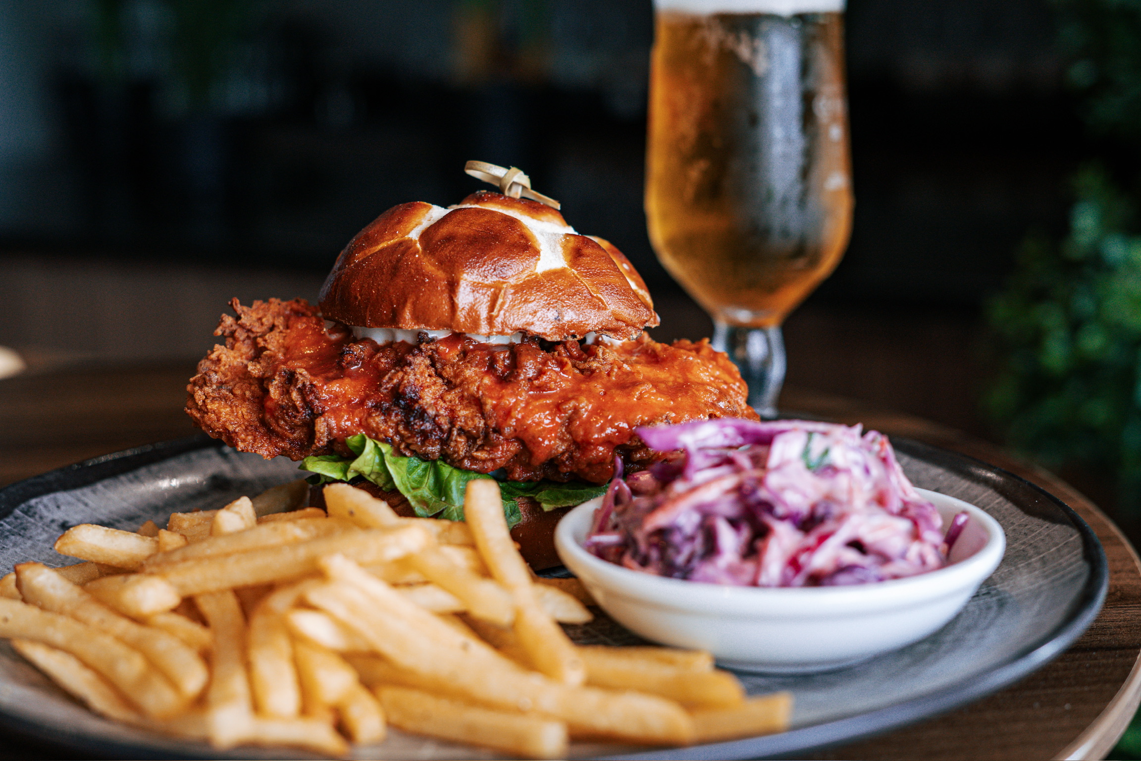 A plate with a fried chicken sandwich on a pretzel bun, a side of fries, and coleslaw. A glass of beer is in the background.