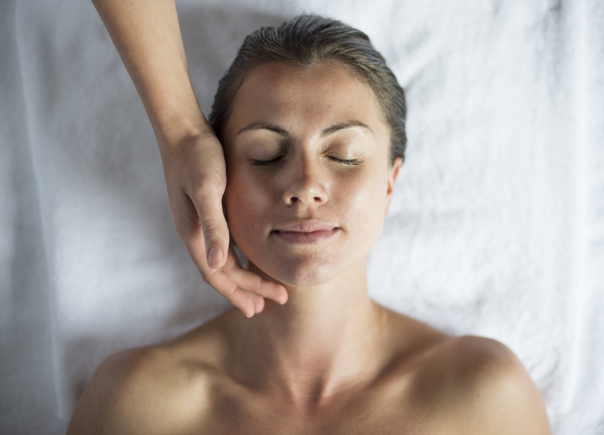 Person lying on a white surface with eyes closed, while a hand rests gently on their cheek.