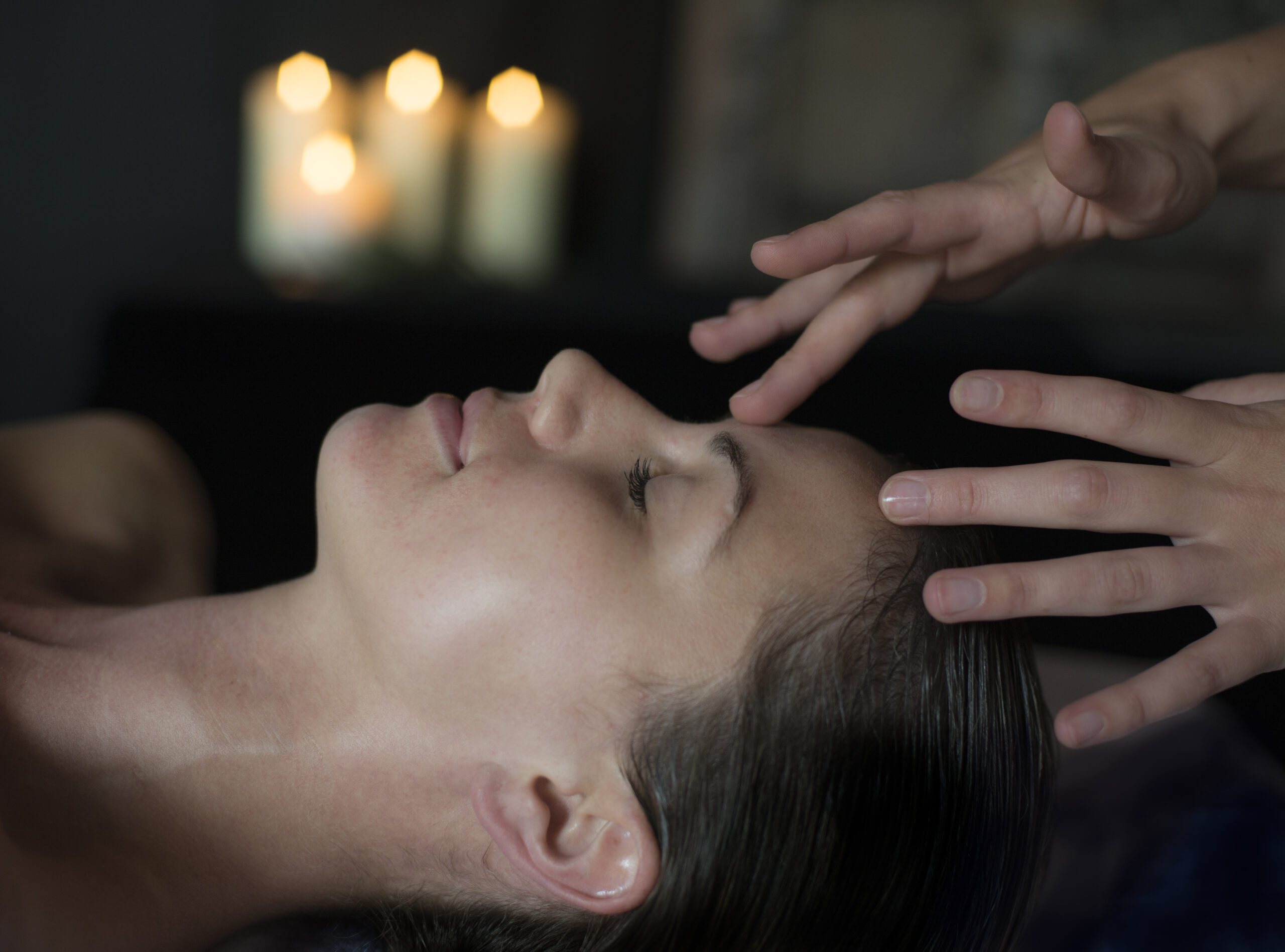 A person lying down with closed eyes as another person's hands hover above their forehead. Three lit candles are blurred in the background.