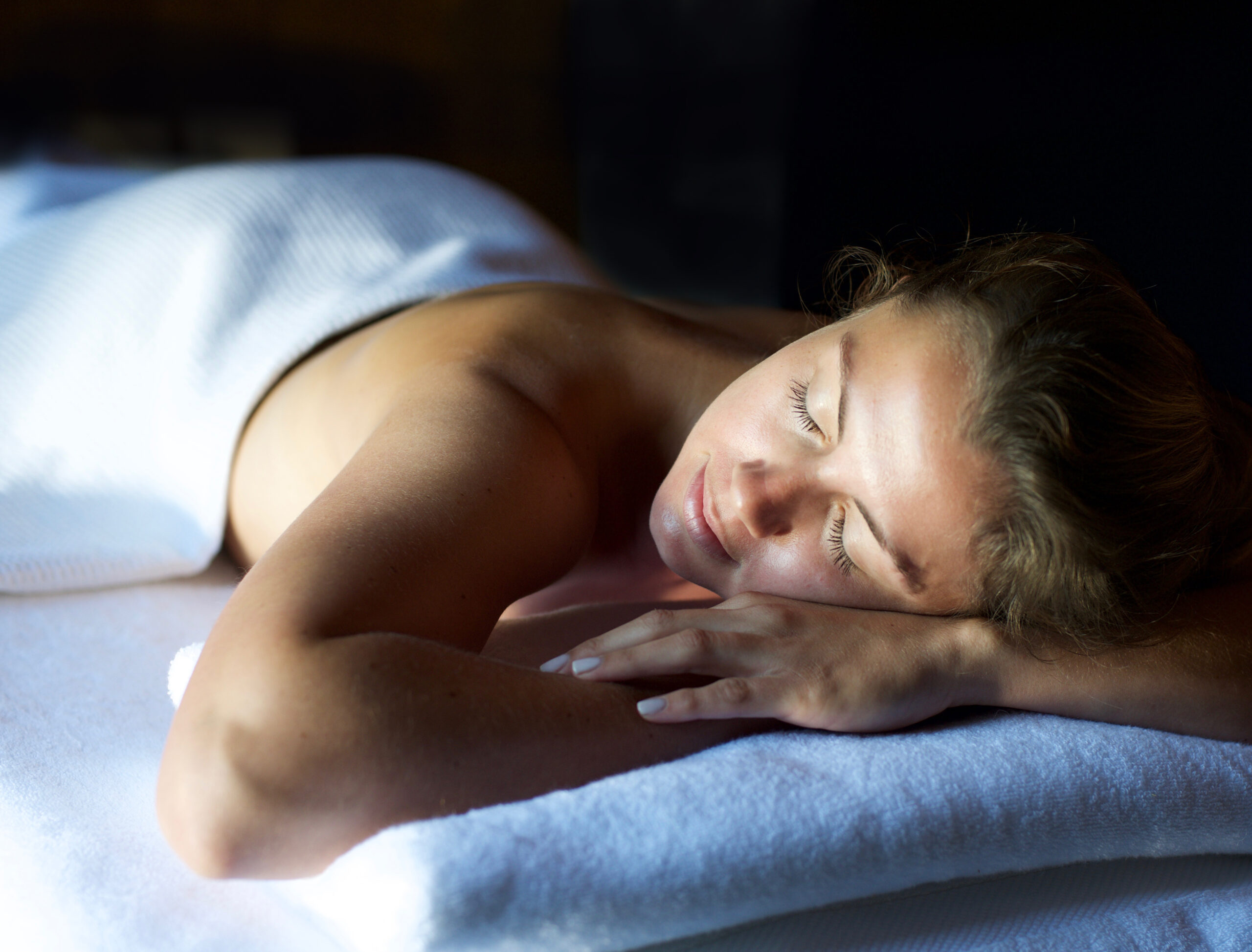 A person is lying on a massage table with eyes closed, partially covered by a white towel, in a relaxed setting with dim lighting.