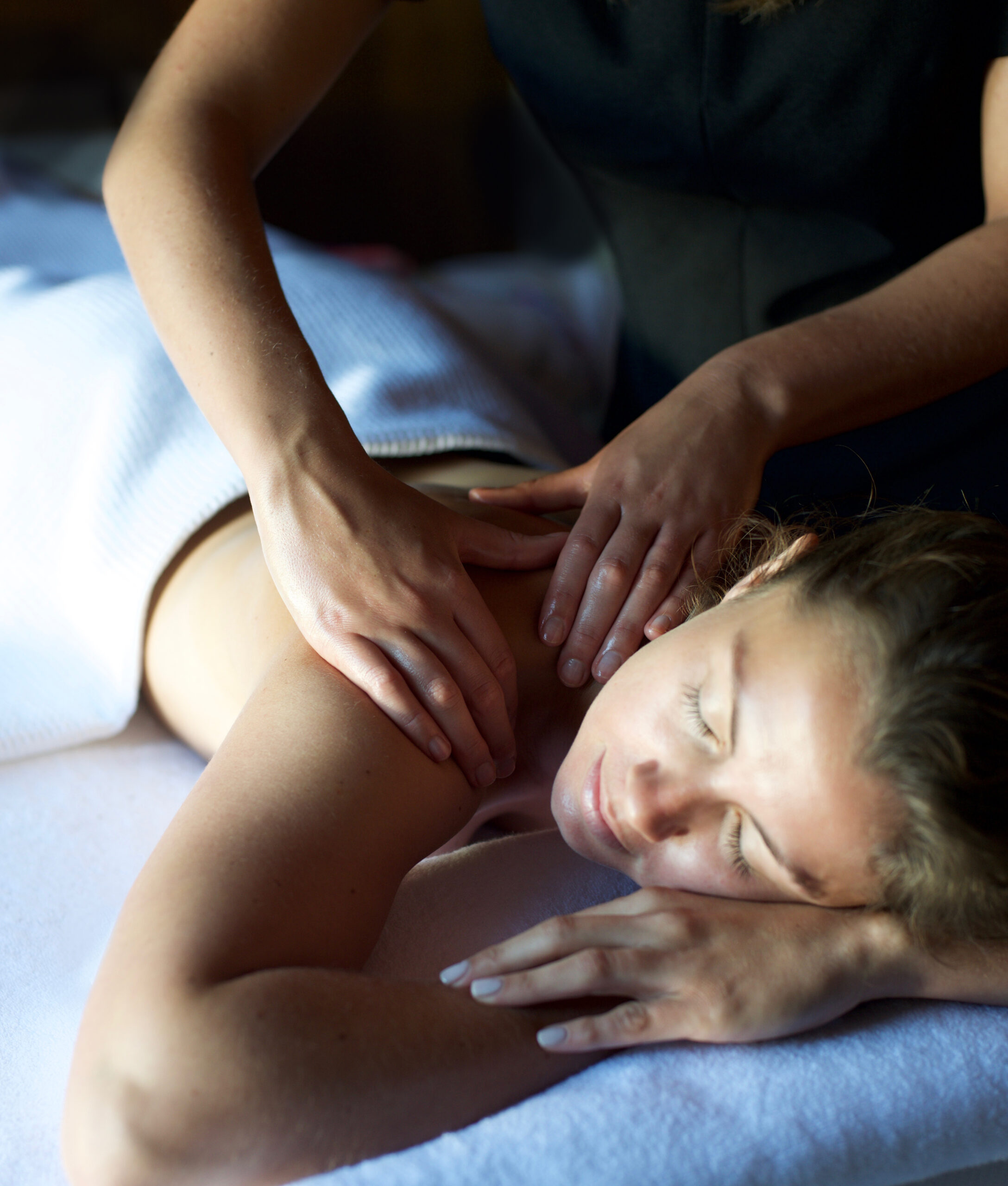 Person receiving a back massage while lying on a white towel, eyes closed, in a relaxed setting.