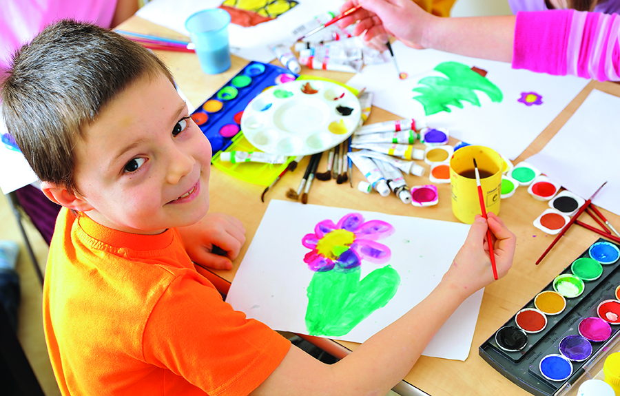 A child in an orange shirt paints a colorful flower with watercolors at a table filled with art supplies.