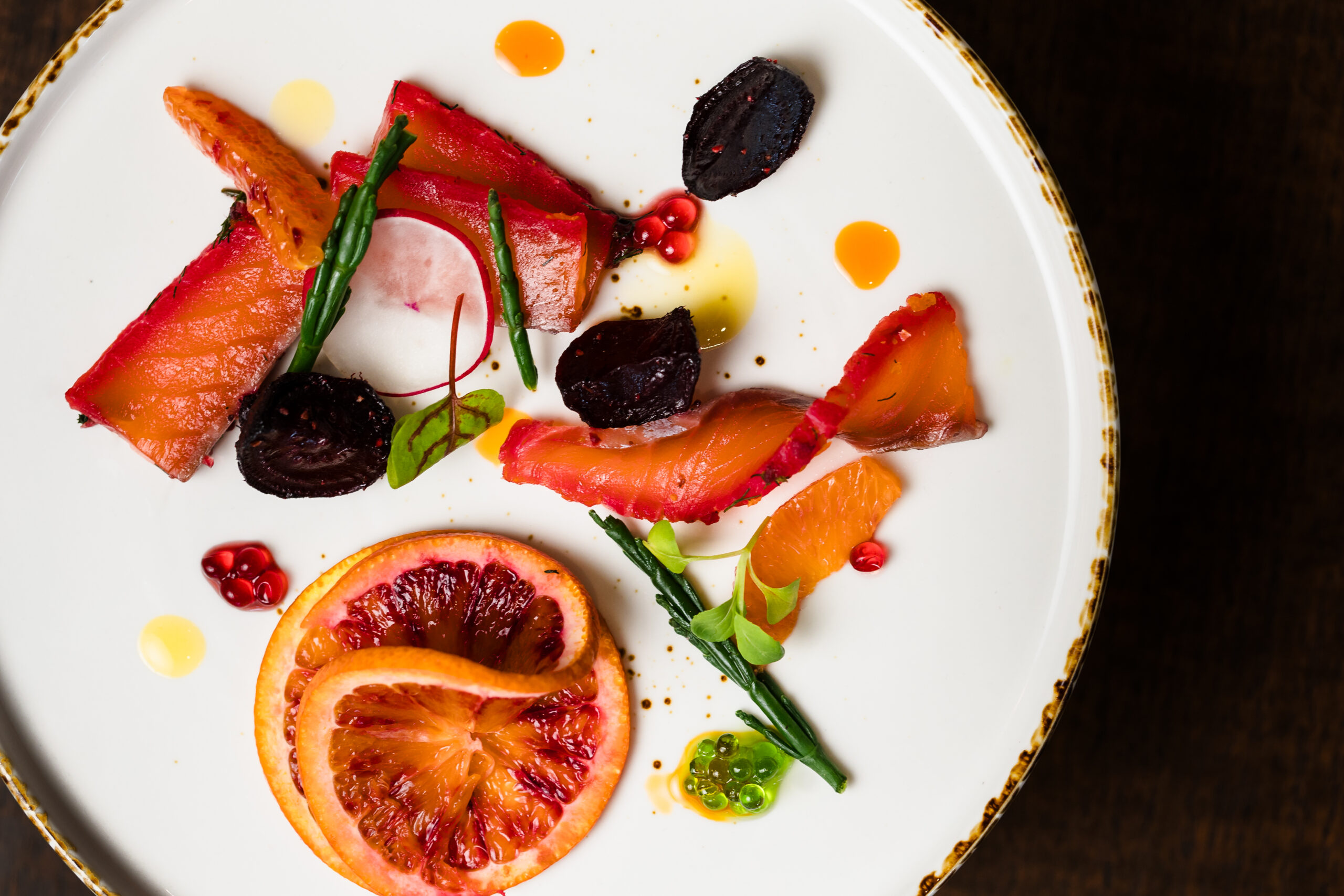 A gourmet dish with sliced fish, orange segments, beets, greens, and a blood orange slice arranged on a white plate.