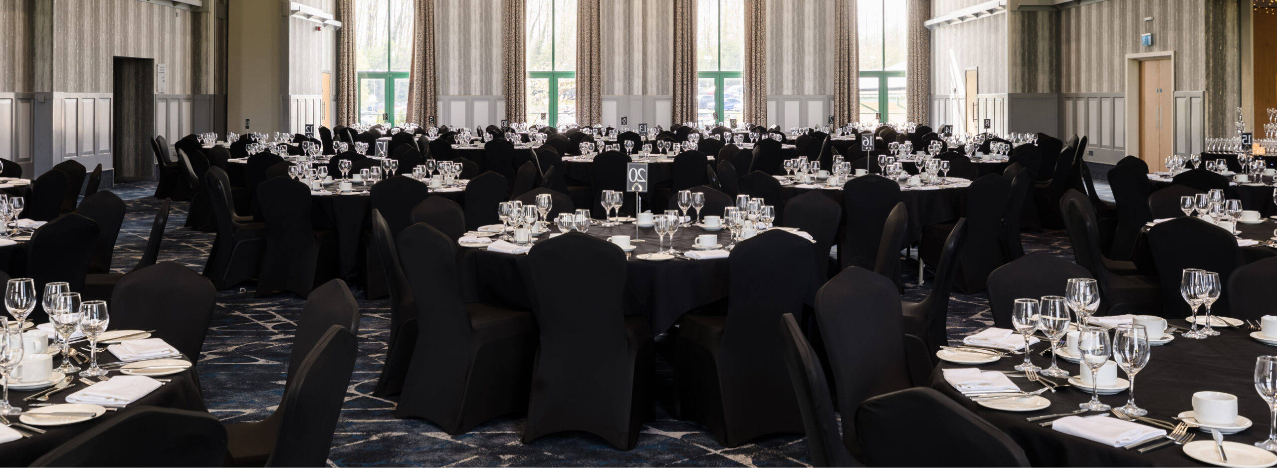 A large banquet hall with round tables set for an event, featuring black chair covers, white tablecloths, and neatly arranged tableware and glasses.