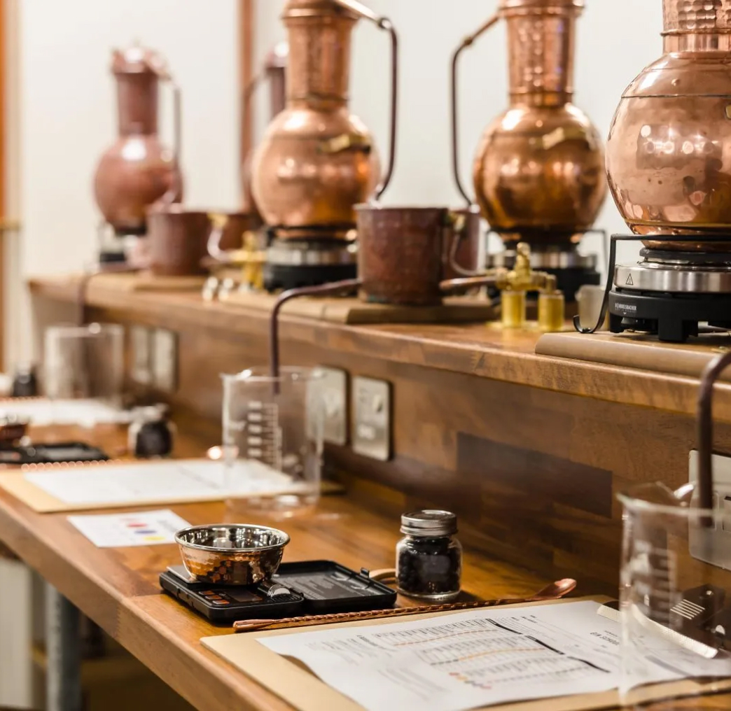 A row of copper stills on a wooden counter with various measuring tools and documents in a distillery setting.