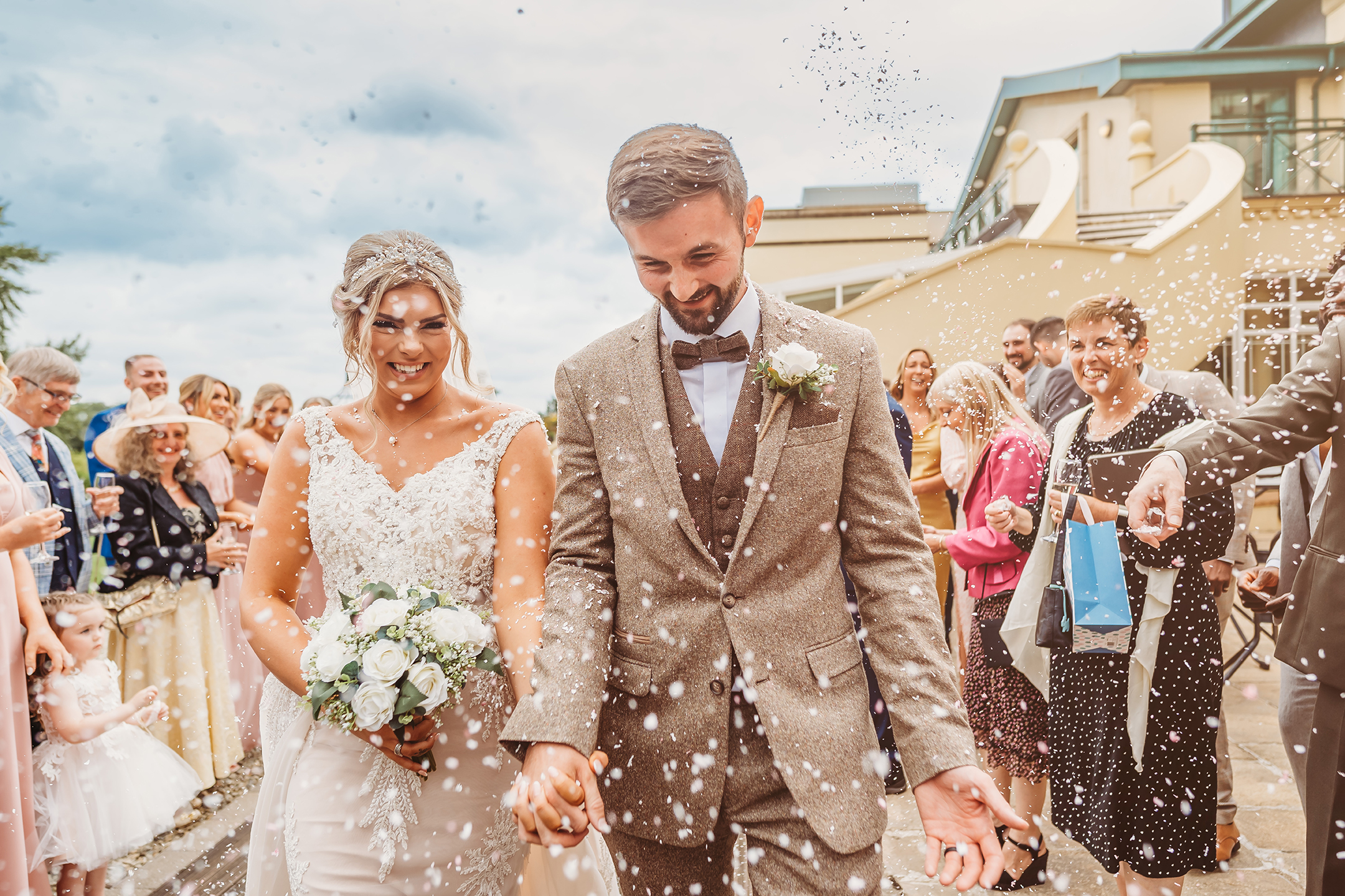 A bride and groom walk hand in hand while guests throw confetti. The bride holds a bouquet, and both are smiling. Other people are visible in the background.