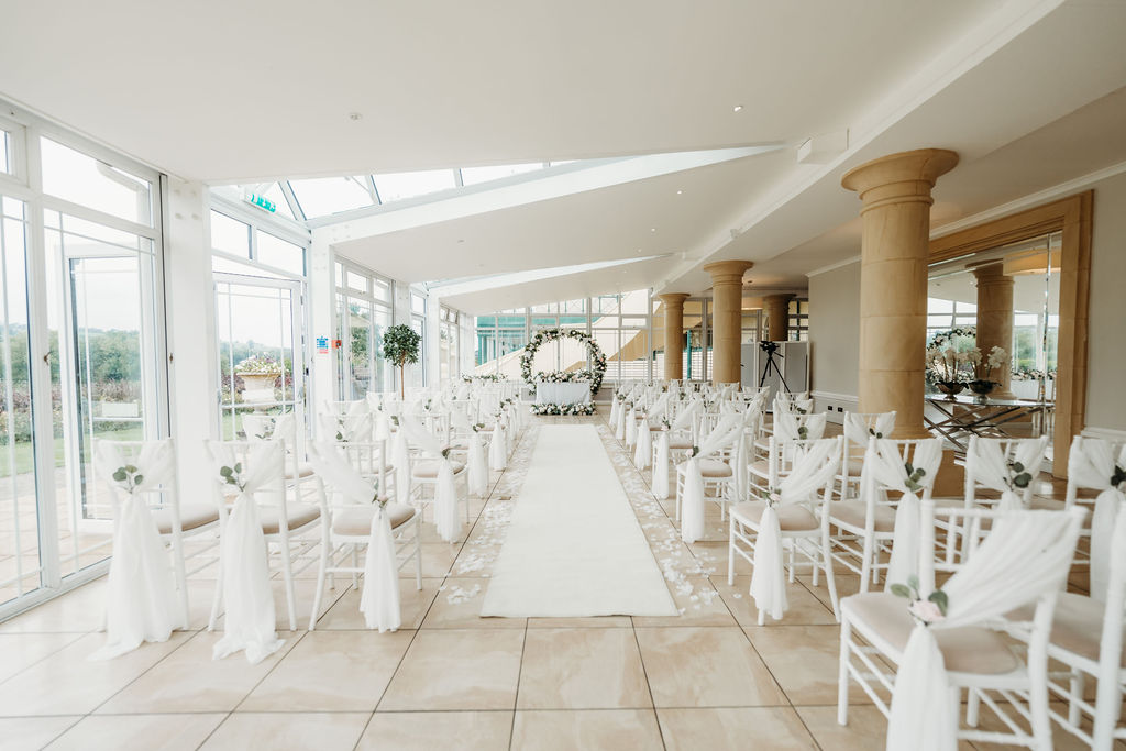 A bright, elegant wedding venue with rows of white chairs and a floral arch at the end of a white aisle runner, set in a spacious room with large windows.