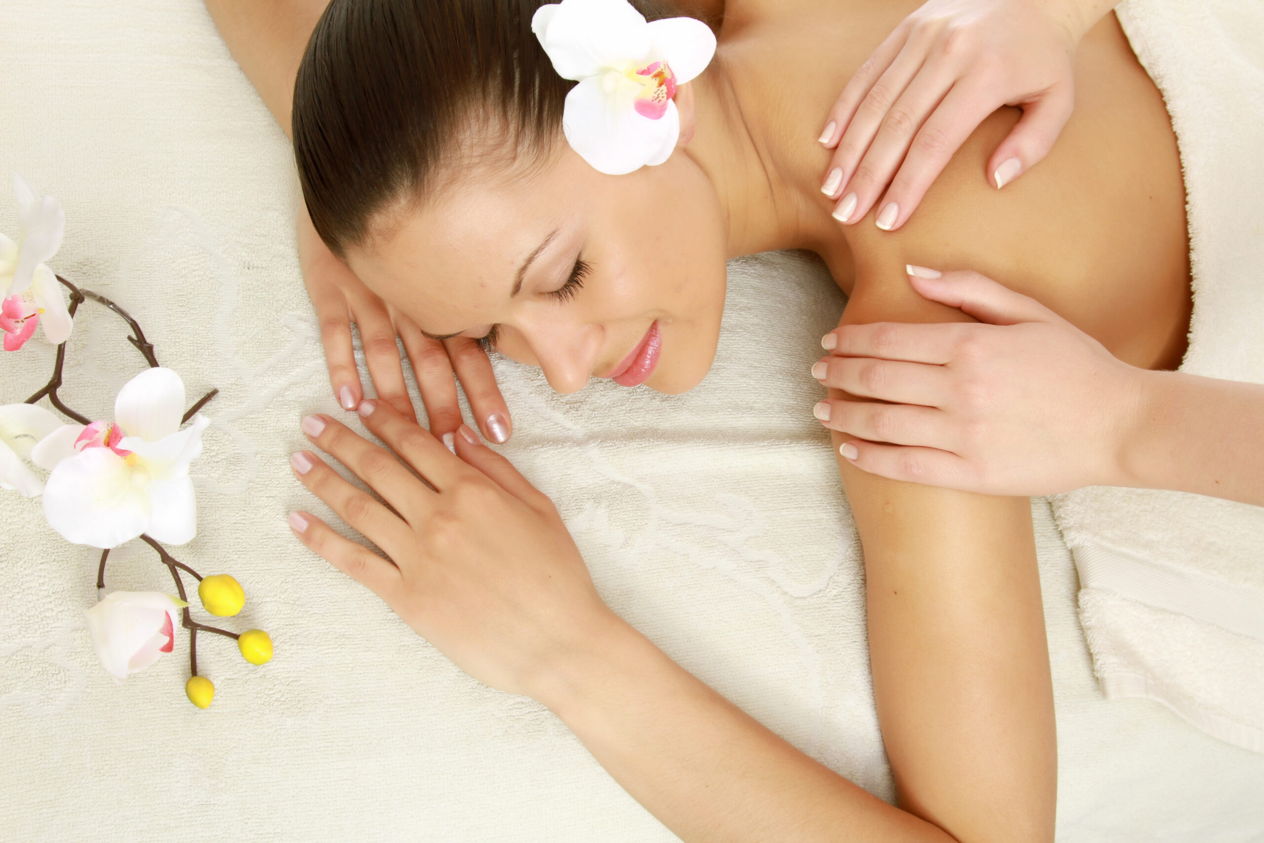 A woman receiving a back massage while lying on a white towel, with an orchid in her hair and yellow buds nearby.