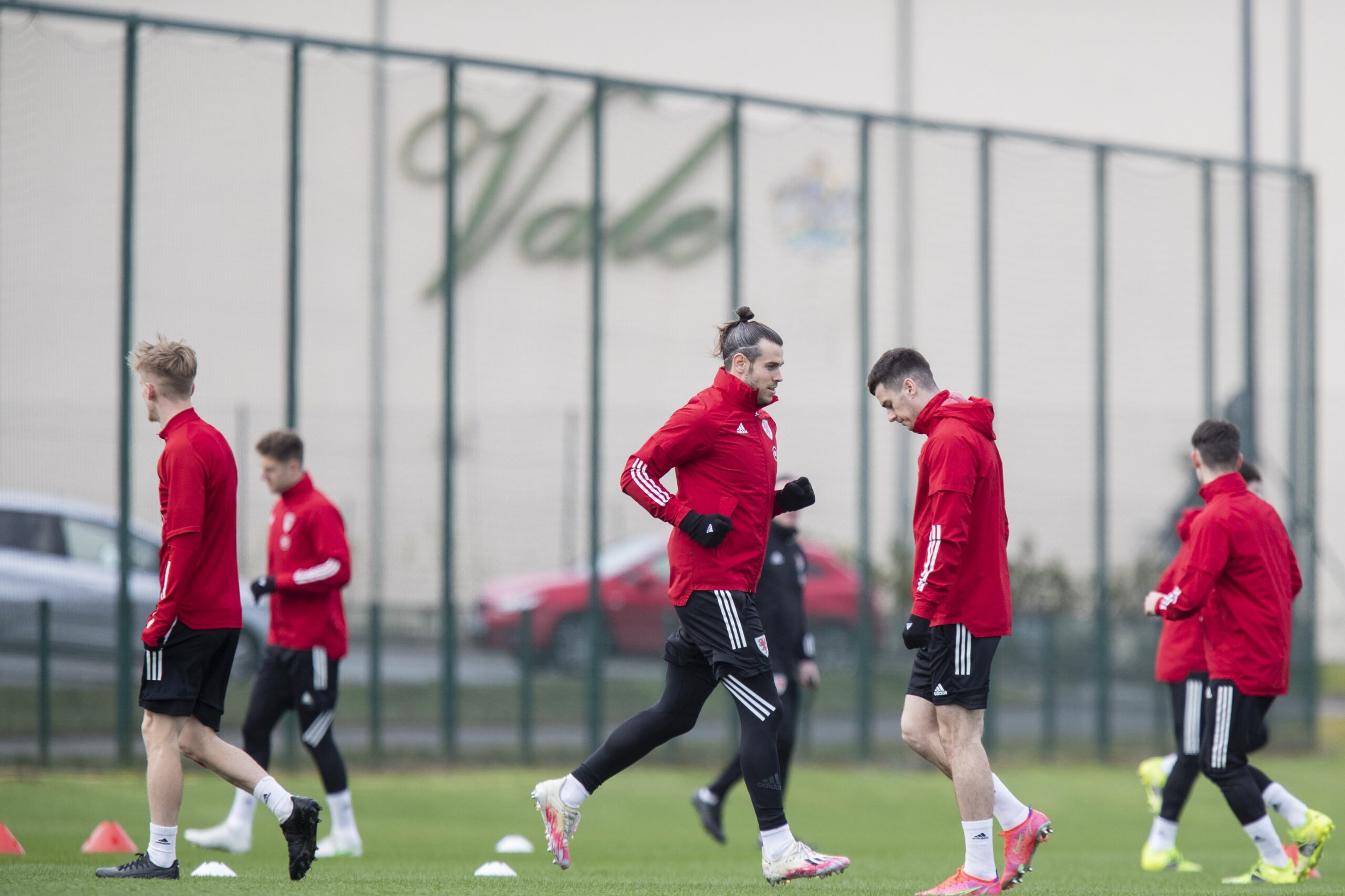 Several football players in red jackets and black pants are training on a grass field with cones, alongside a fence.