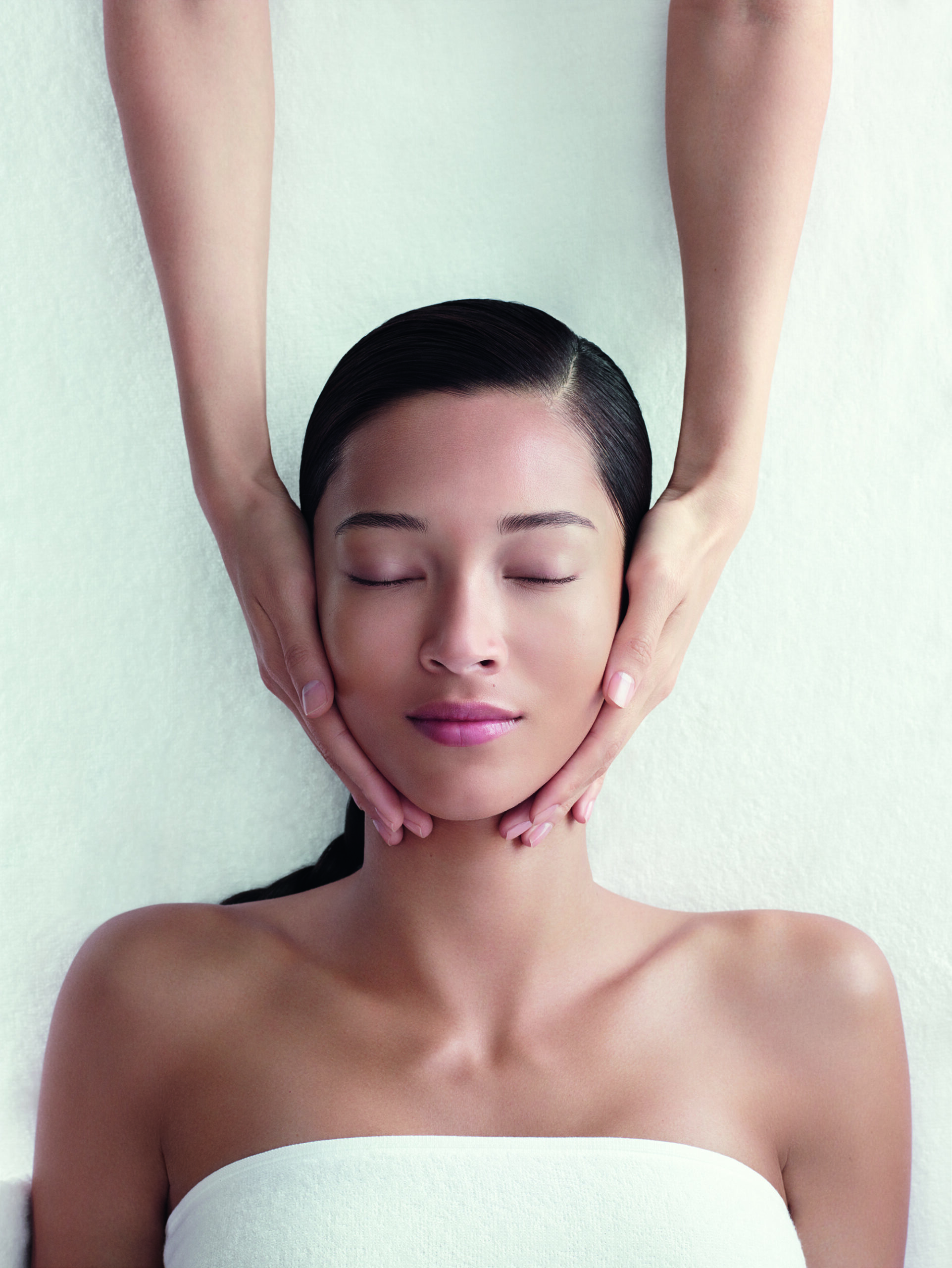 A person with eyes closed receives a facial massage while lying on a white surface. Hands are gently placed on their cheeks.