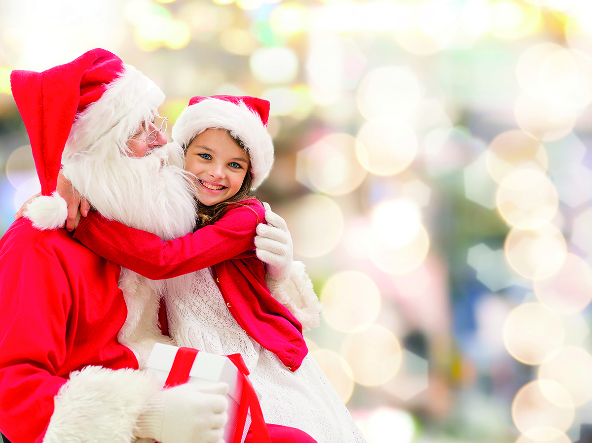 A child in a Santa hat hugs a person dressed as Santa Claus, holding a gift. The background is blurred with festive lights.