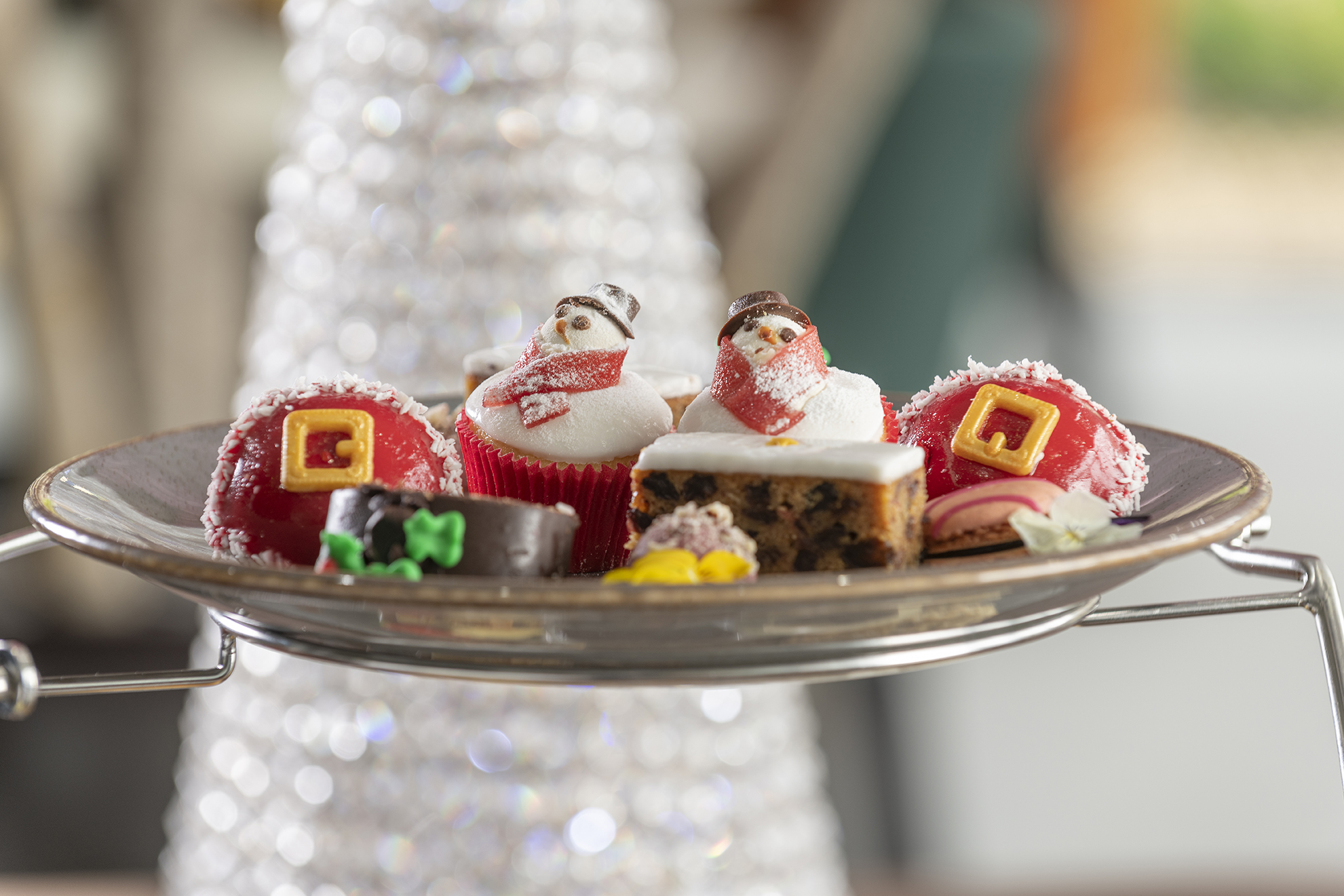 A festive dessert platter with cupcakes, Christmas-themed mousse, and assorted holiday sweets, displayed in front of a blurred white Christmas tree.