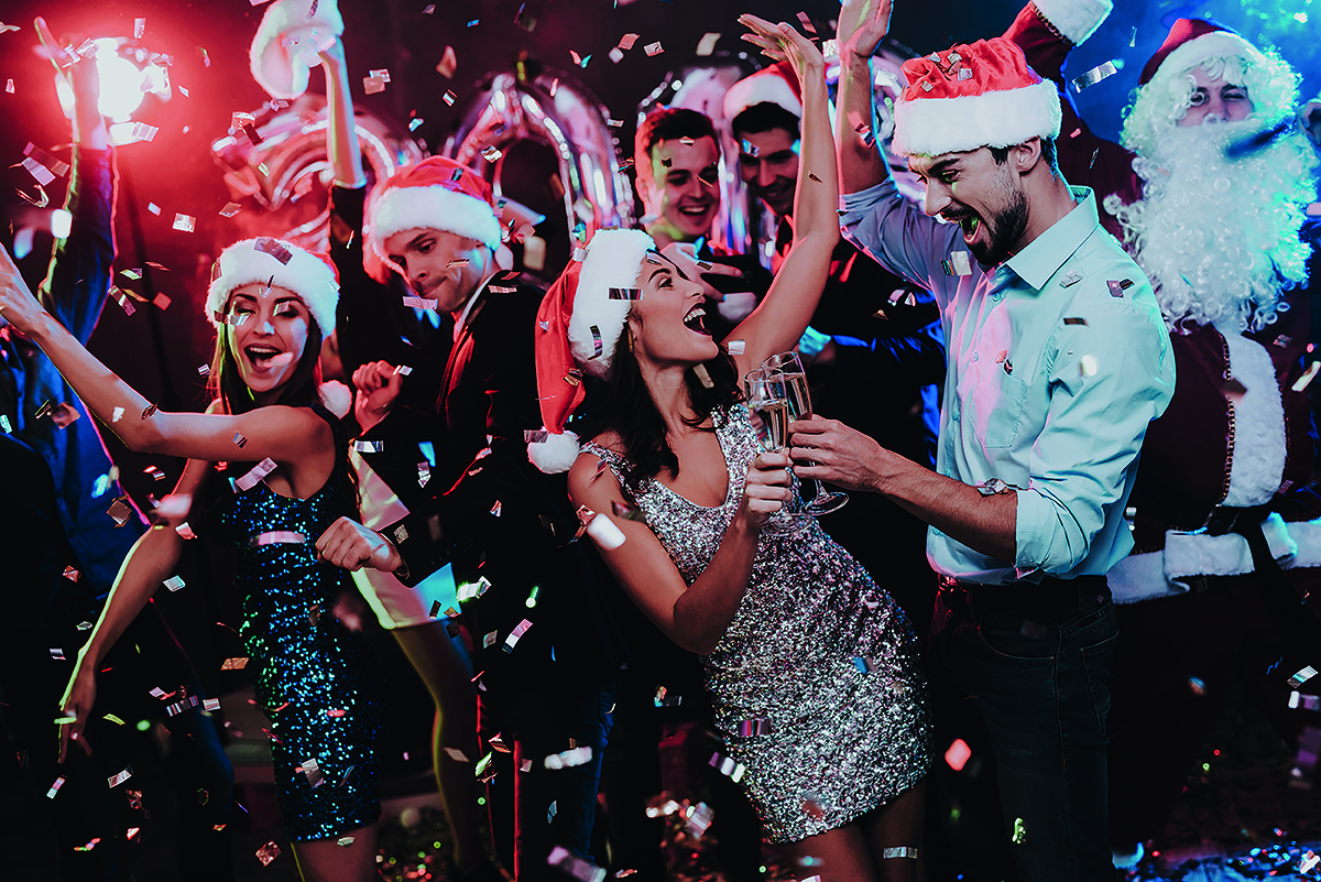 People wearing Santa hats and festive outfits celebrate at a lively party with confetti and champagne.