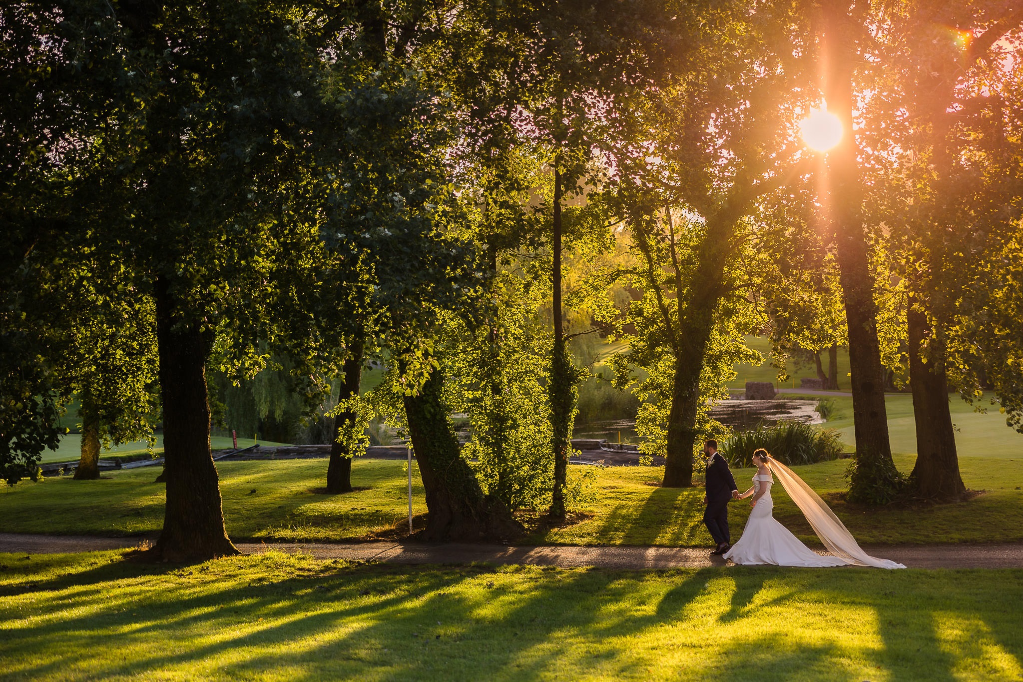 A bride and groom walk along a tree-lined path bathed in warm sunlight, with long shadows cast on the grass.