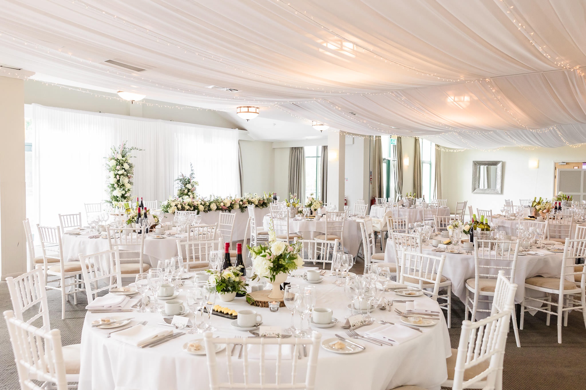 A decorated wedding reception hall with round tables covered in white linens, floral centerpieces, and white chairs. The ceiling is draped with white fabric and string lights.