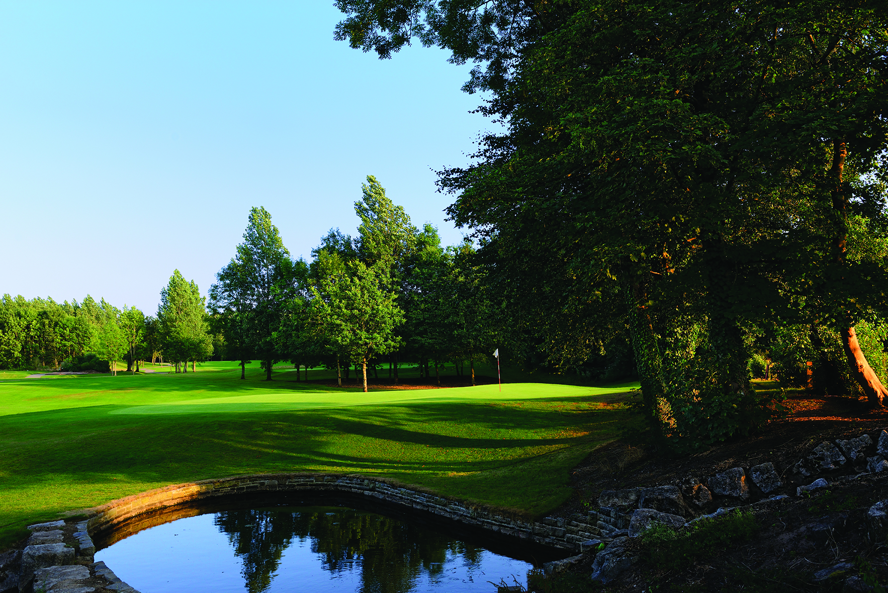 A peaceful golf course with lush green fairways, bordered by tall trees, under a clear blue sky. A small pond with a stone edge is visible in the foreground.