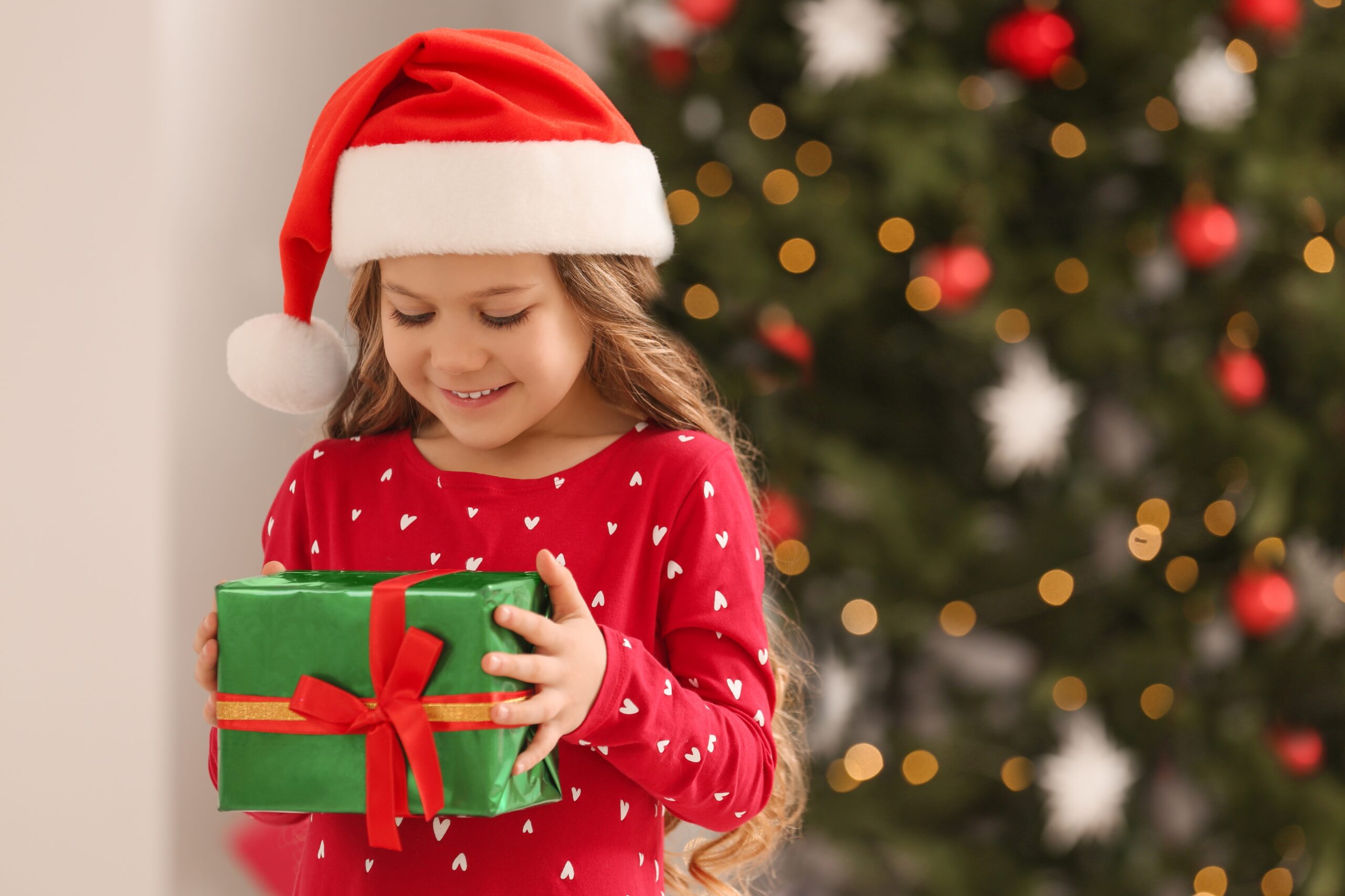 Young girl in a Santa hat holding a gift with red ribbon, standing in front of a decorated Christmas tree.