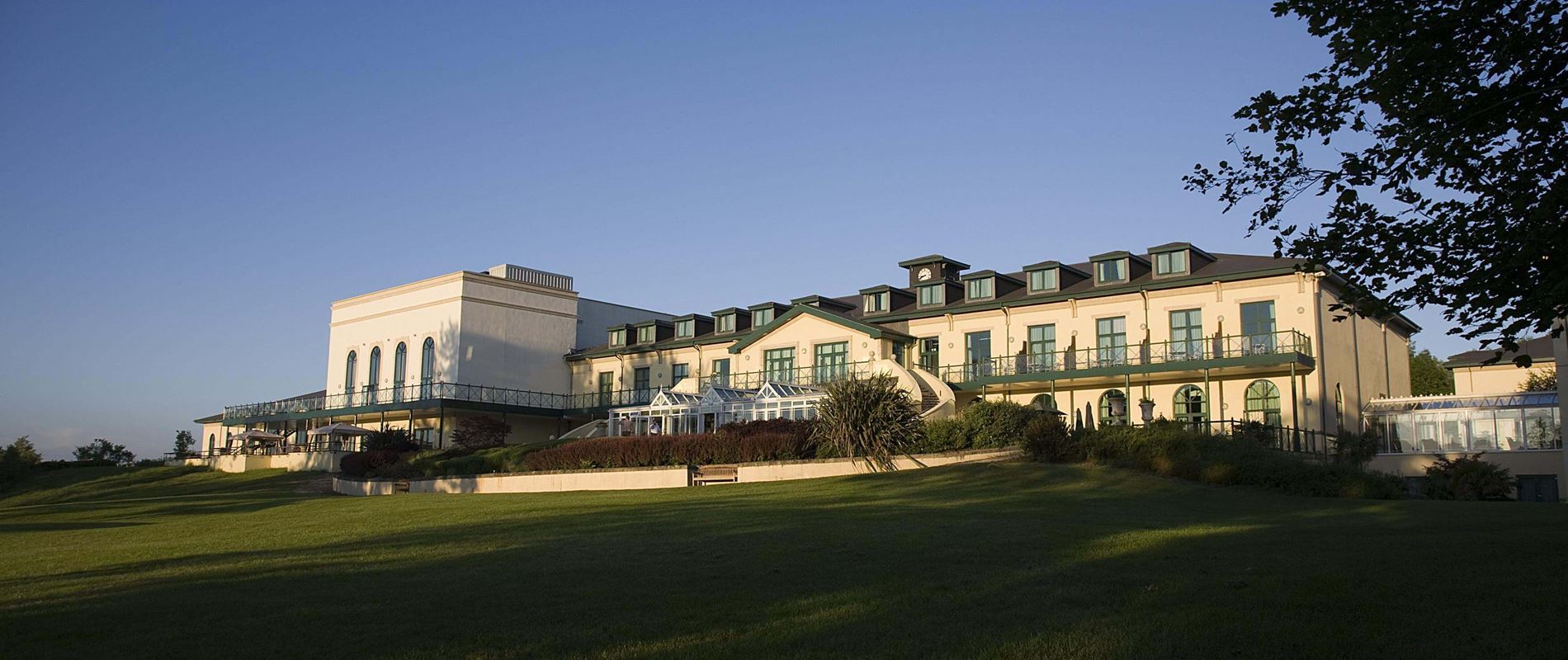 Large beige hotel building with balconies and green trim, set on a grassy landscape under a clear blue sky.