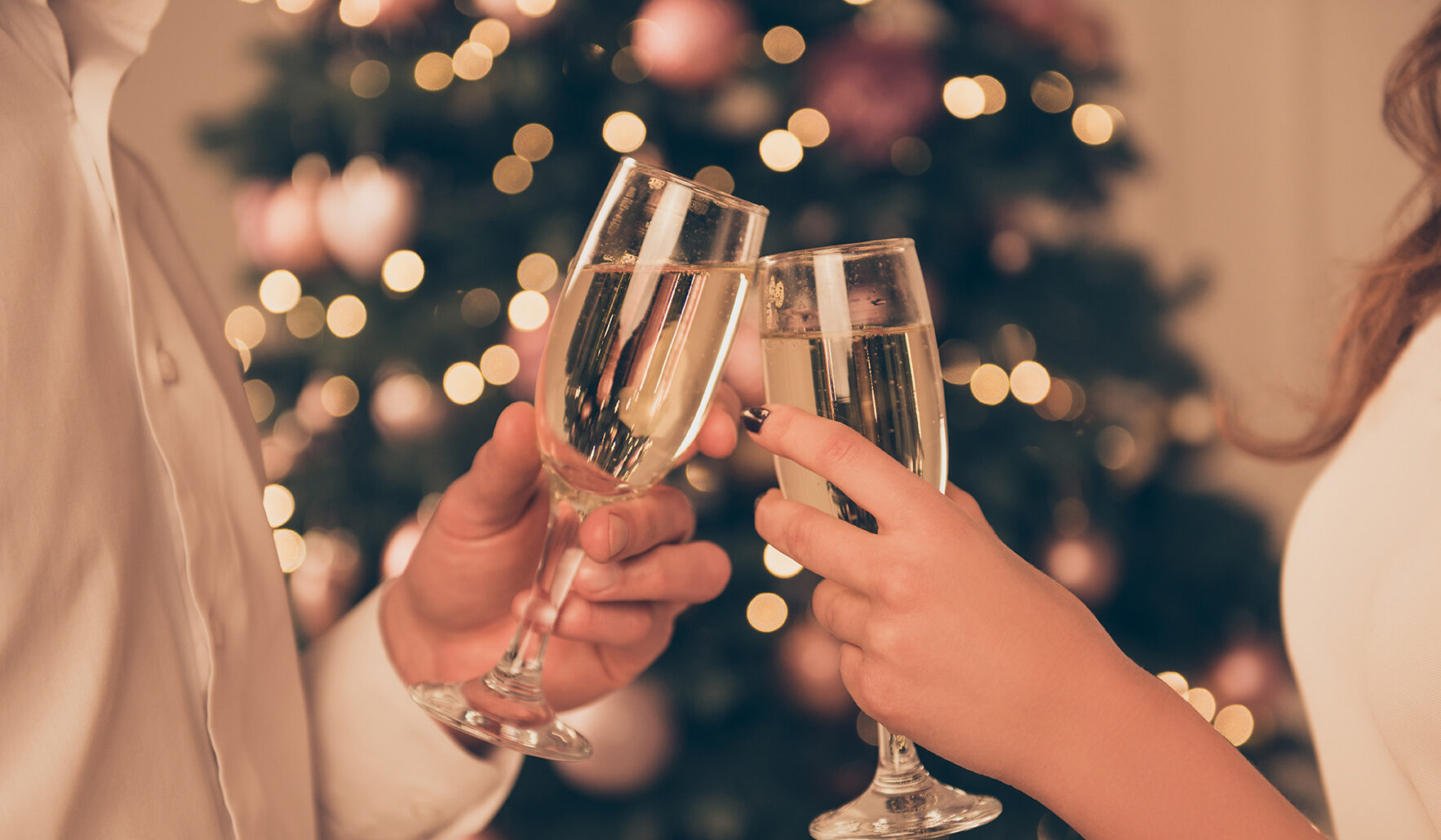 Two people clinking champagne glasses in front of a decorated Christmas tree.