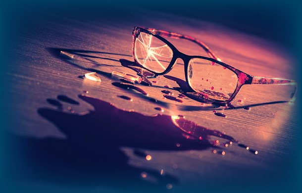 Broken glasses with a cracked lens are lying on a surface next to a pool of liquid, possibly wine or juice, under dramatic lighting.