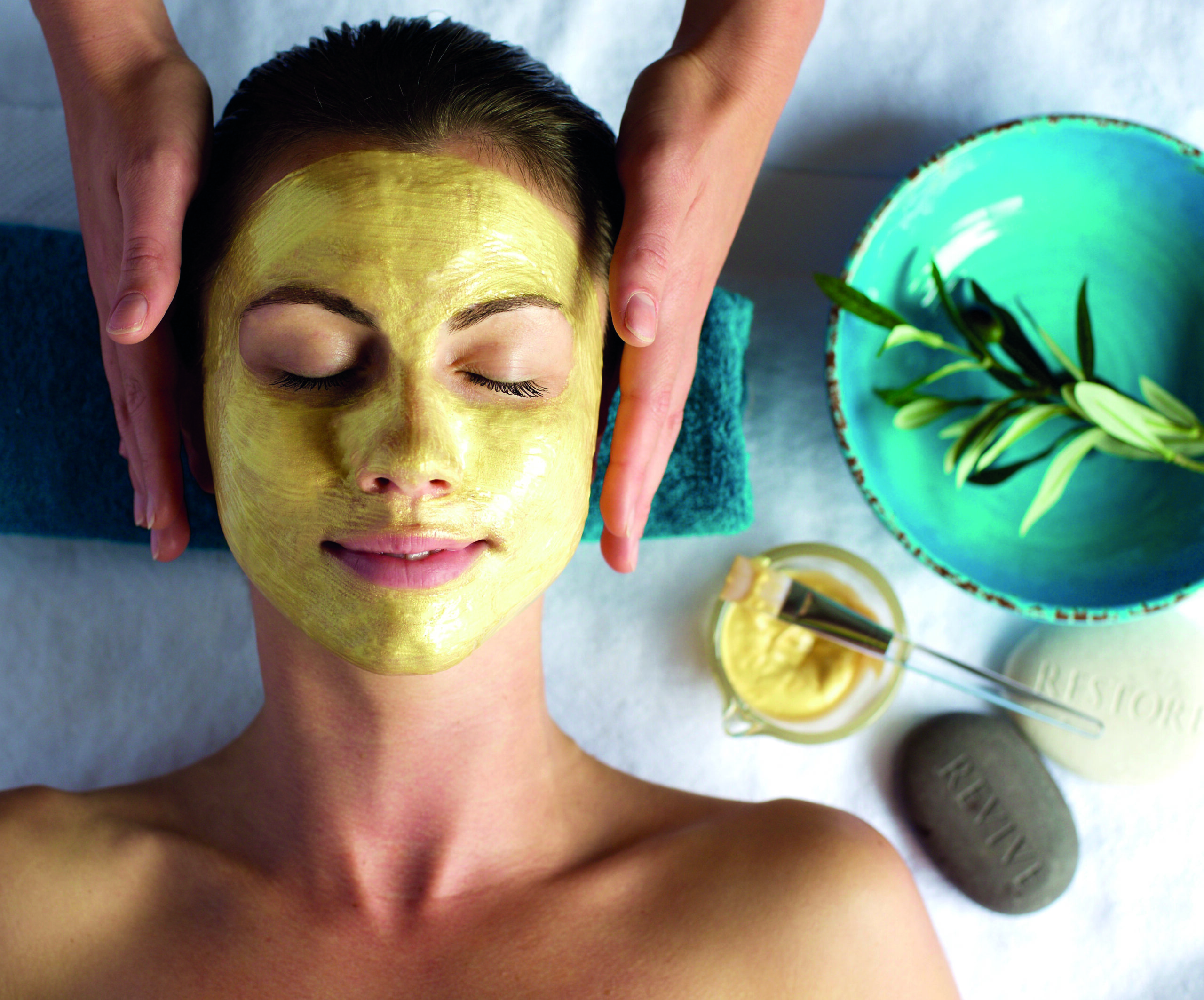 A person receiving a facial treatment with a yellow mask applied, lying on a towel. Nearby are a bowl with leaves, a dish with cream, a stone labeled 