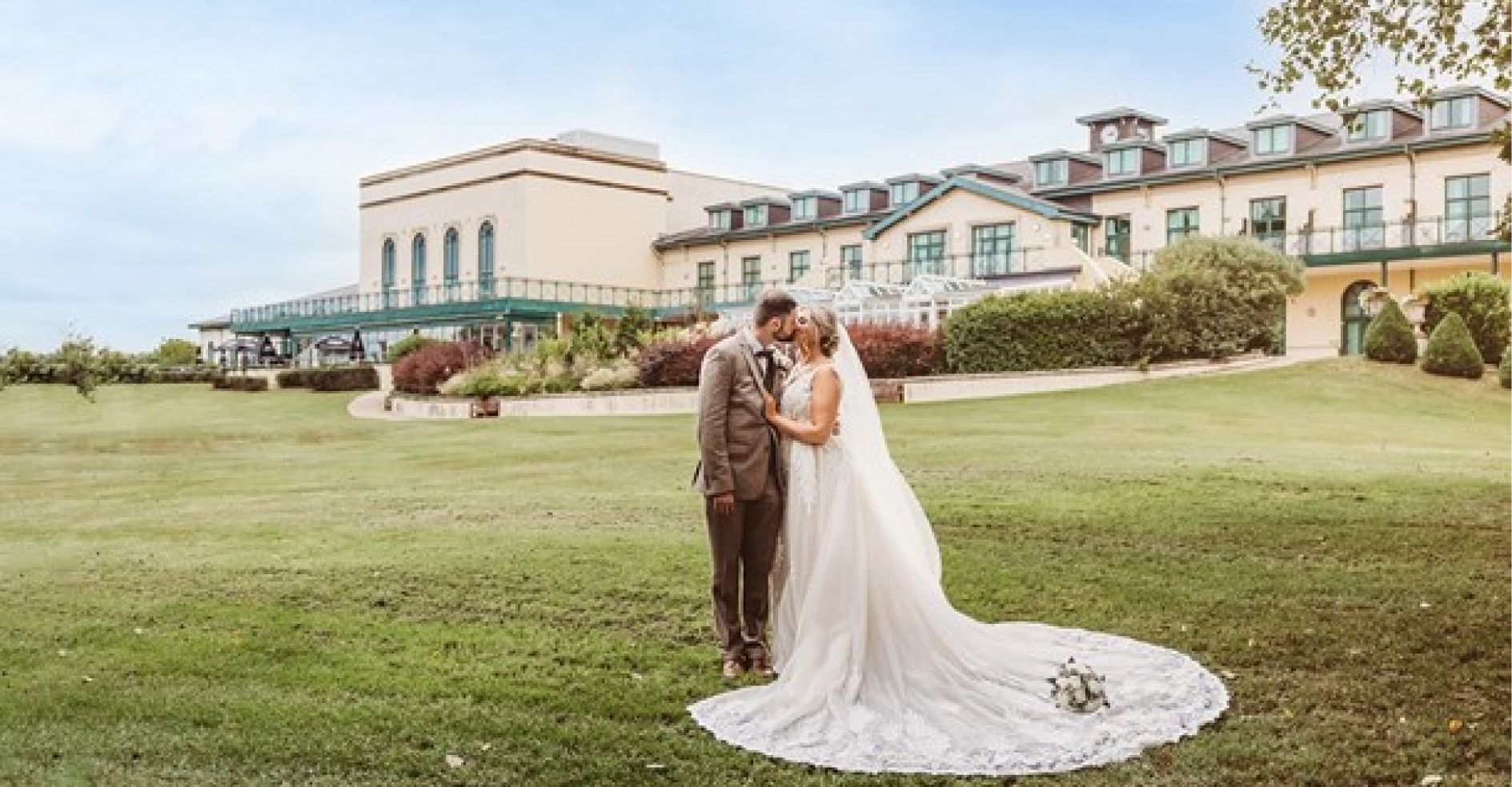 A bride and groom stand together on a grassy lawn in front of a large building. The bride wears a long white gown with a train, and the groom is in a suit. A bouquet lies on the ground.