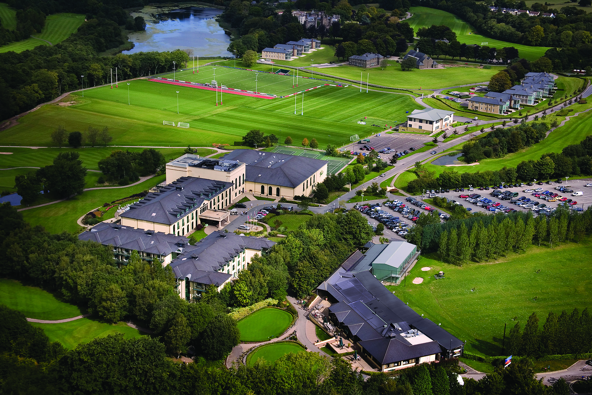 Aerial view of a sports complex with multiple buildings, green fields, rugby pitches, parking lots, and surrounding trees.