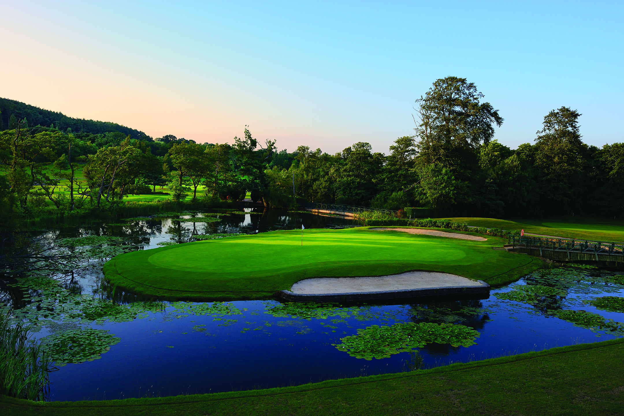 A golf course with a green surrounded by water and trees under a clear blue sky.