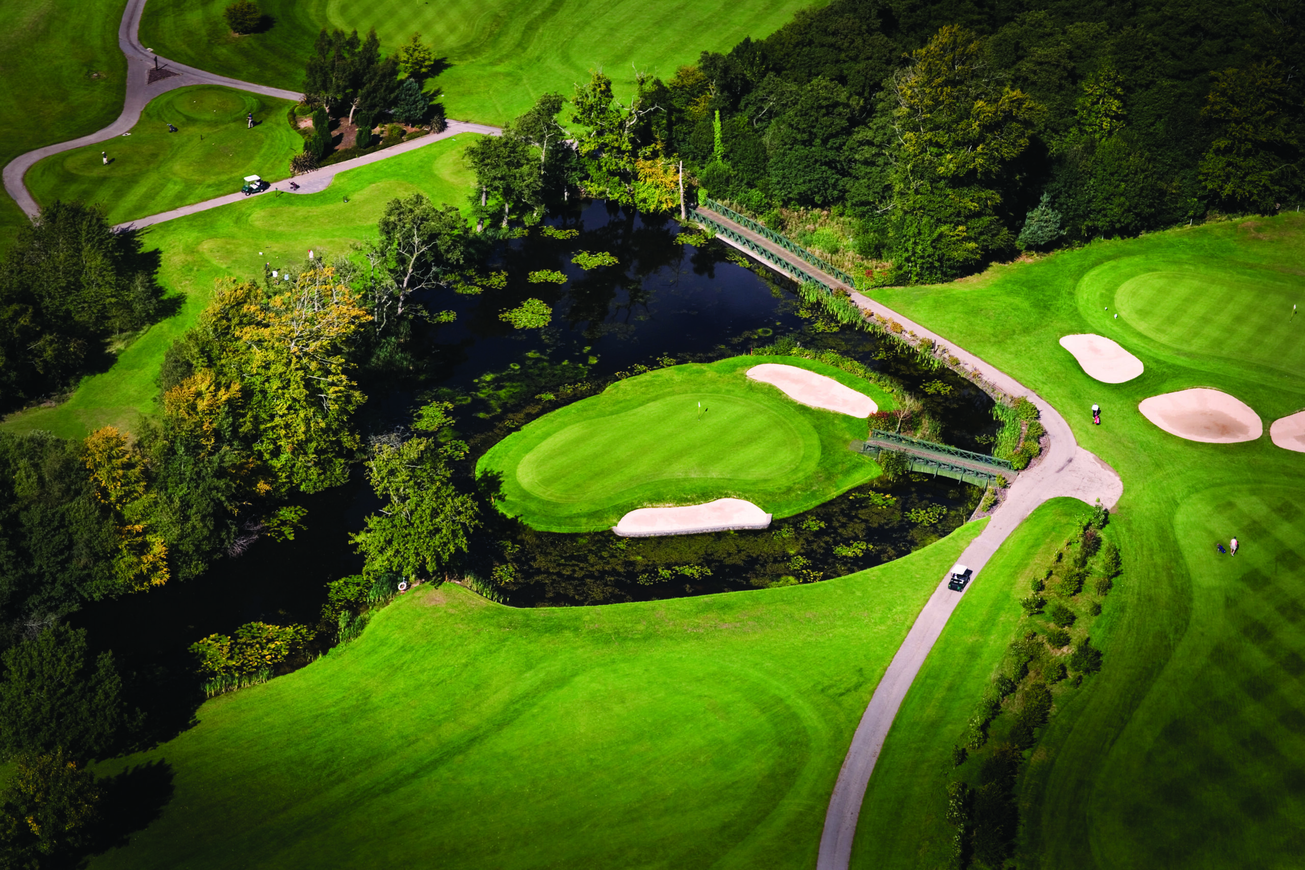 Aerial view of a golf course with a small lake, surrounding greenery, sand bunkers, and a pathway.