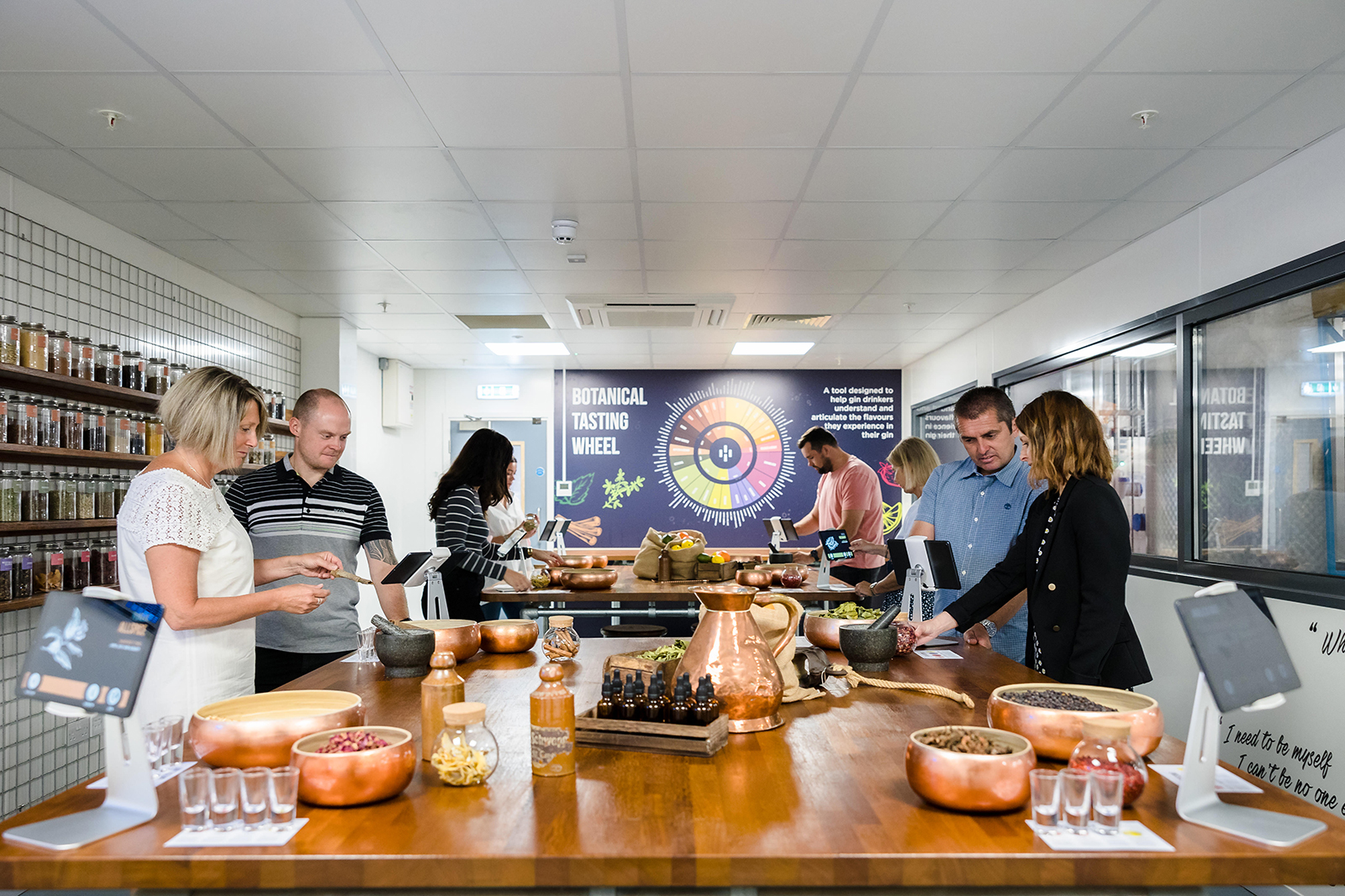 People participating in a botanical tasting event, examining various ingredients on a wooden table, with informational displays in the background.