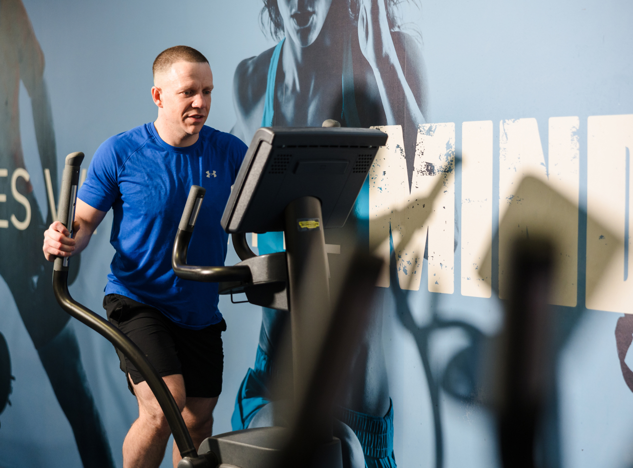 Person in a blue shirt using an elliptical machine at the gym with a motivational wall graphic in the background.