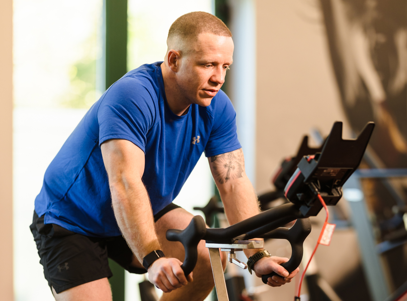 Man in a blue shirt and black shorts using a stationary bike at a gym, focused on the screen in front.