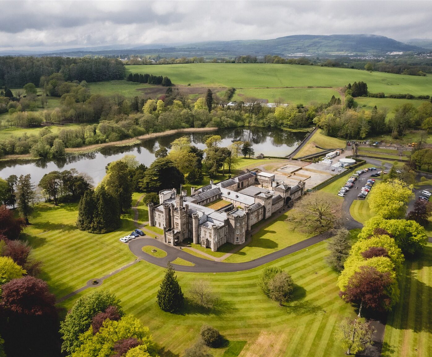 Aerial view of a large historic building surrounded by green lawns, trees, and a lake, with parked cars along a driveway.