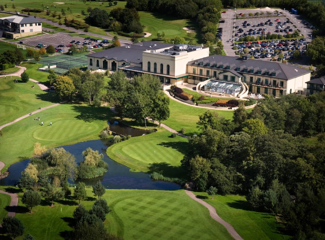 Aerial view of a golf resort with a clubhouse, parking lot, landscaped gardens, golf course, ponds, and surrounding trees.