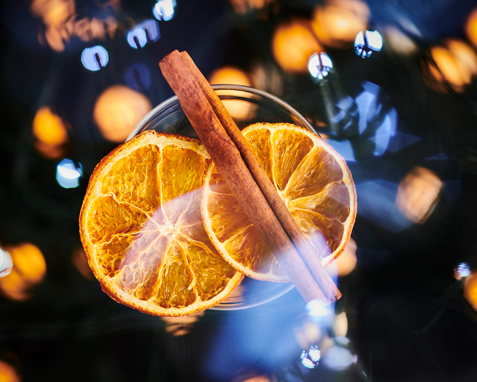 A glass with dried orange slices and cinnamon sticks, set against a background of blurred lights.