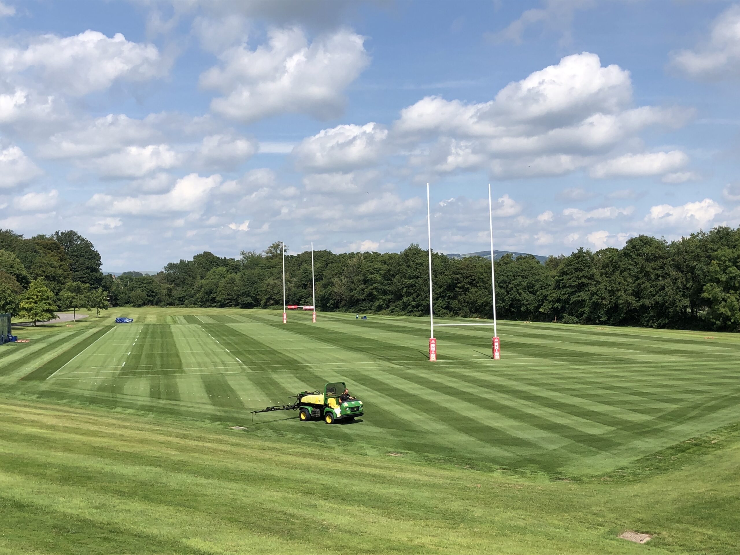 A tractor mowing grass on a striped rugby field under a partly cloudy sky, with goalposts visible in the distance.