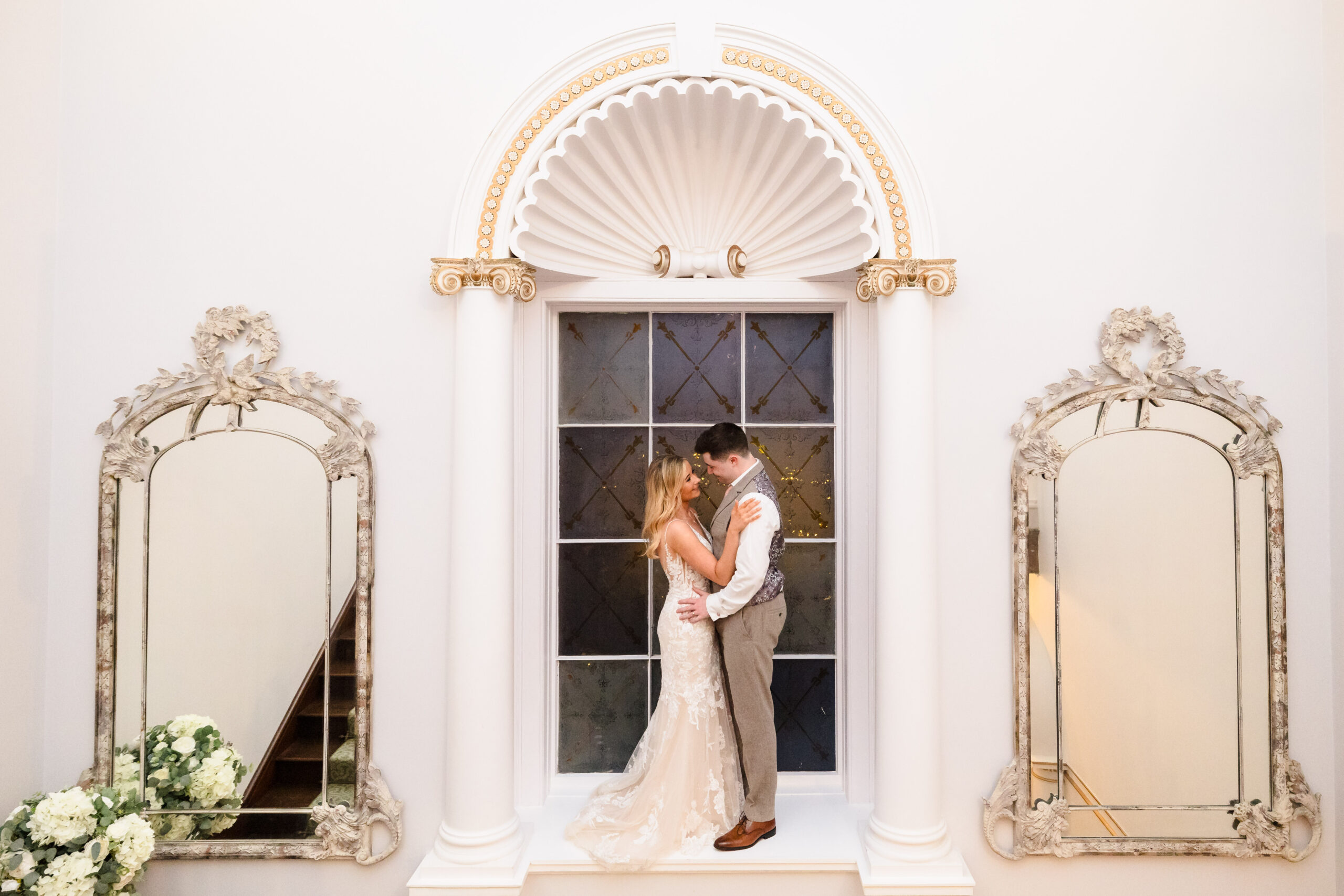 A couple embraces in front of an ornate window, flanked by two decorative mirrors. The setting is elegant and brightly lit.