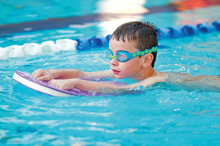 A young boy wearing goggles practices swimming with a kickboard in a pool.