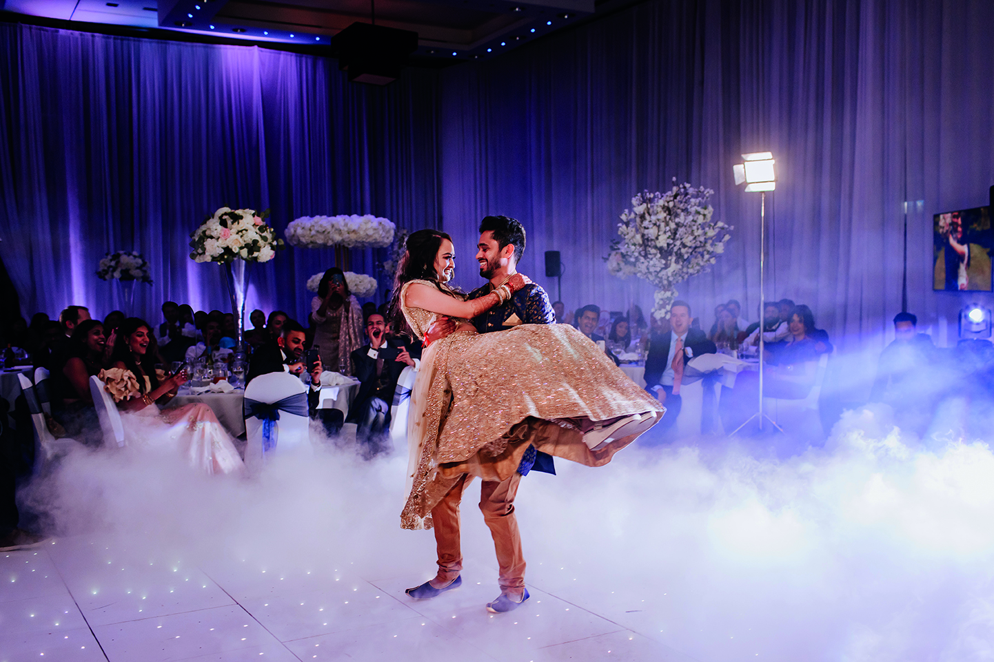 A man lifts a woman while dancing at a formal event with guests seated around. The room is lit in blue tones with floral decorations and fog effects on the dance floor.