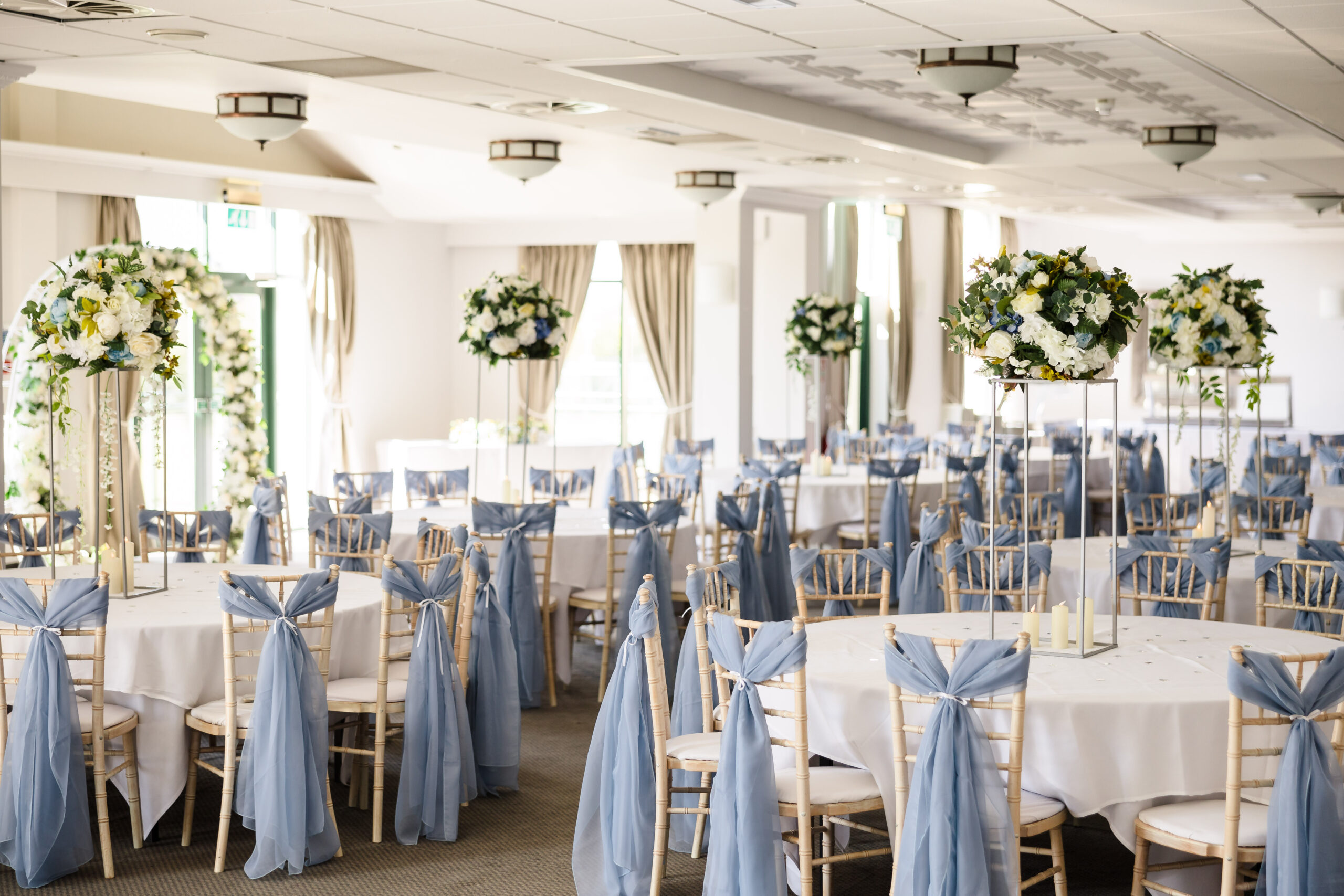 A decorated wedding reception hall with round tables, white tablecloths, and chairs draped with blue fabric. Tall floral centerpieces adorn the tables.