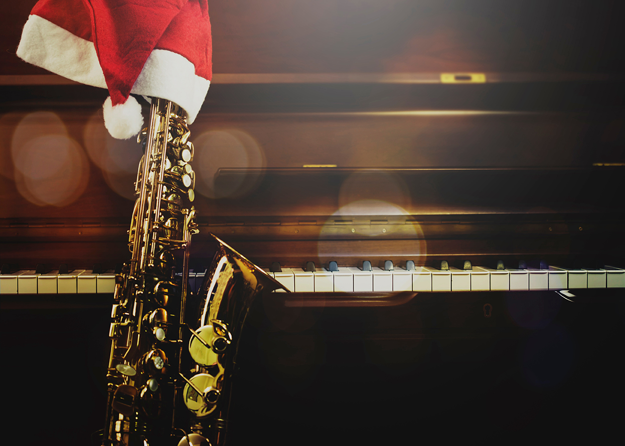 A saxophone with a Santa hat rests against a piano keyboard, with blurred lights in the background.