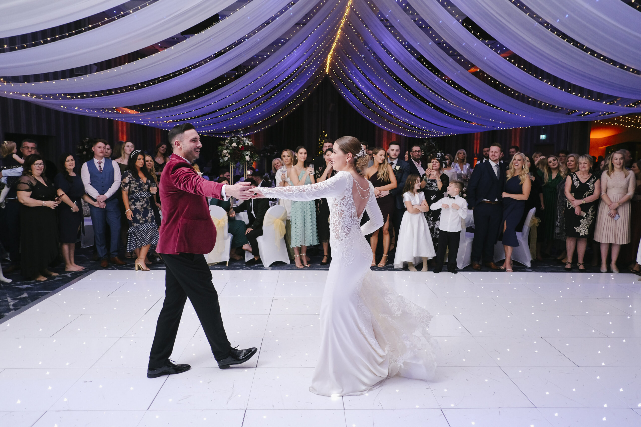 A couple dances in the center of a large, decorated hall, surrounded by an audience. Draped lights hang from the ceiling.