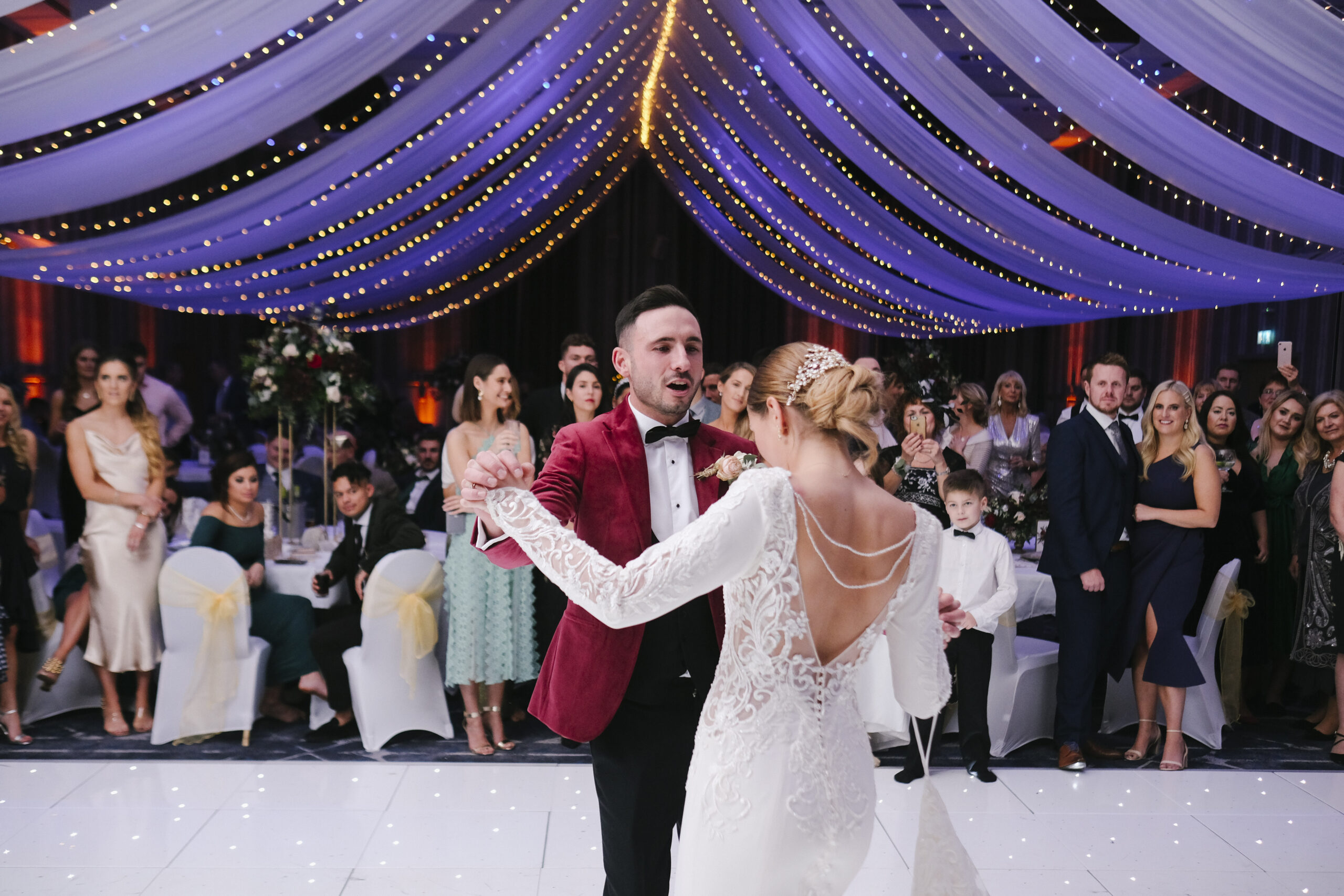 A couple dances under draped fabric and string lights at a formal event, surrounded by seated and standing guests.